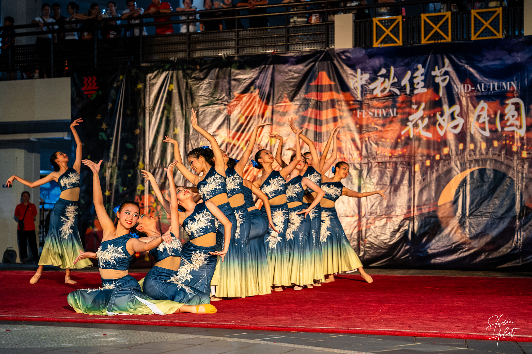 Dance troupe in blue dresses performs on red carpeted stage, arms raised, with audience balcony above.