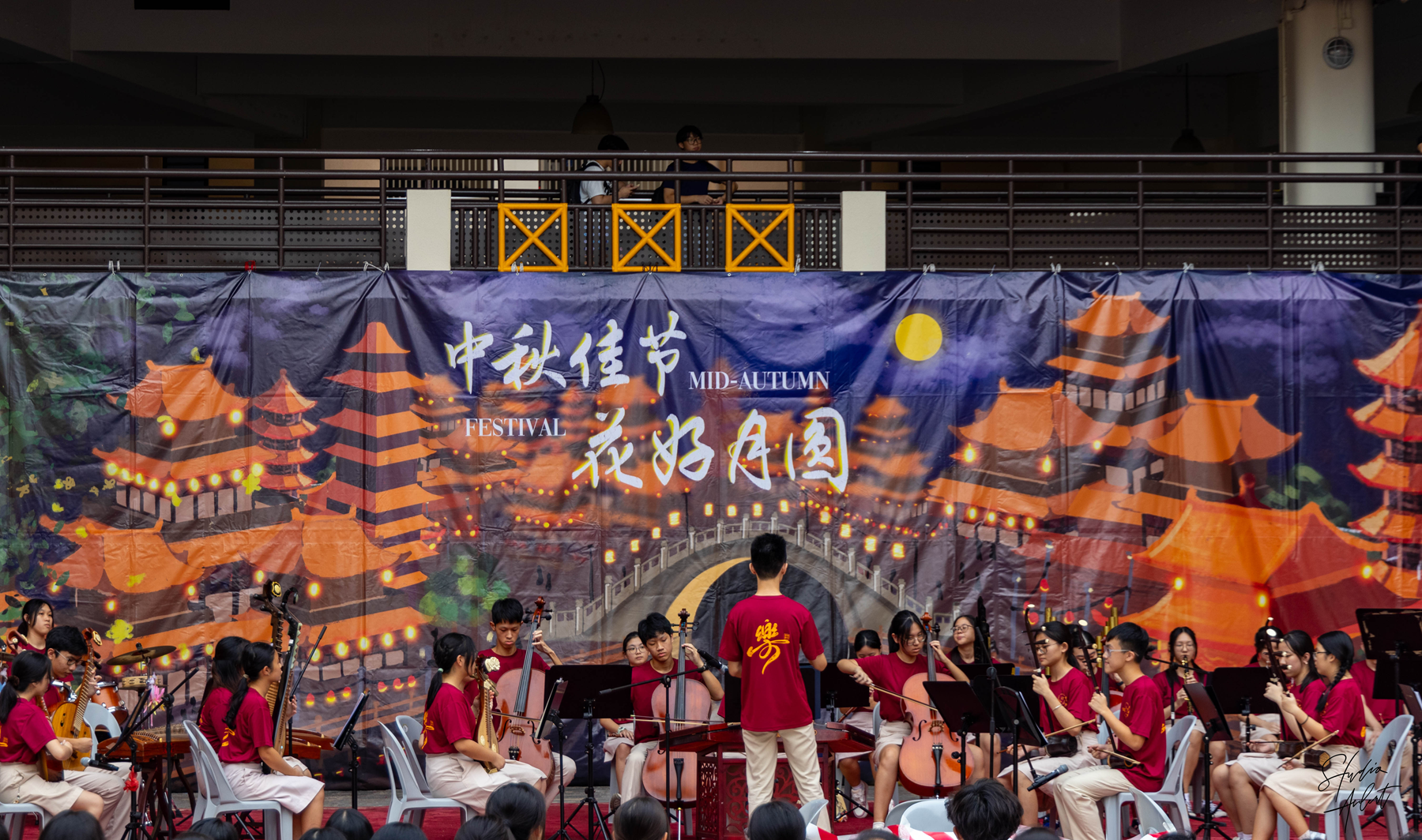 Youth orchestra in red shirts performs at a Mid-Autumn Festival event before a colorful Chinese-themed banner.