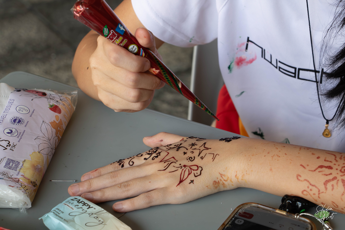 A close-up shot of a student intricately applying a dark henna design onto the back of another student's hand using a cone dispenser.