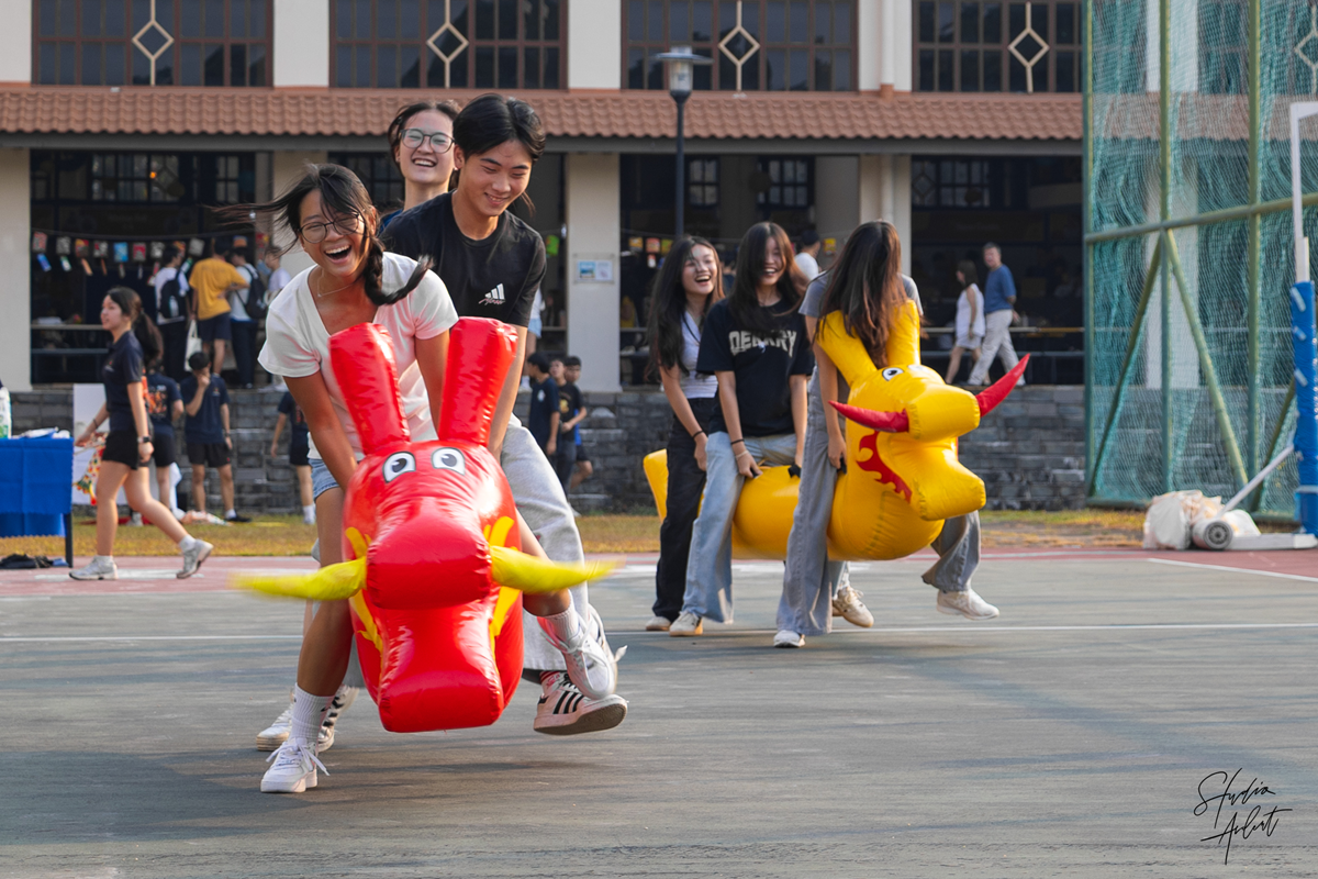 Students laughing while competing in an inflatable dragon boat race game on an outdoor campus court during the Mid-Autumn Festival.