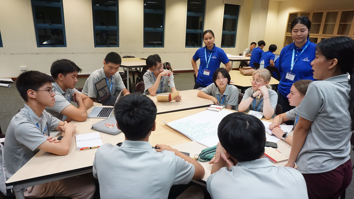A group of students sitting around a round table engaged in a collaborative discussion with laptops and notebooks open.