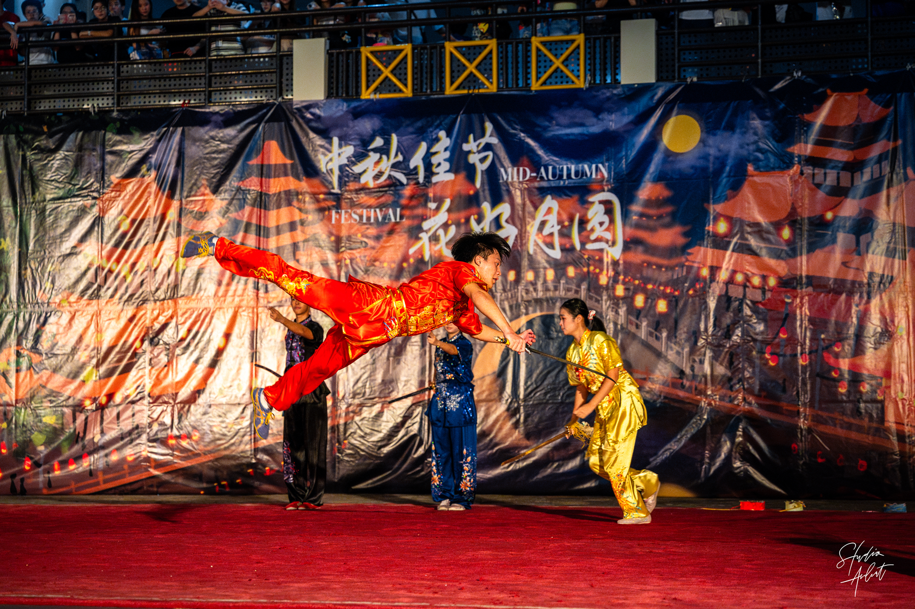 Martial artist in red midair kick while two performers in blue and gold hold staffs on a festival stage.