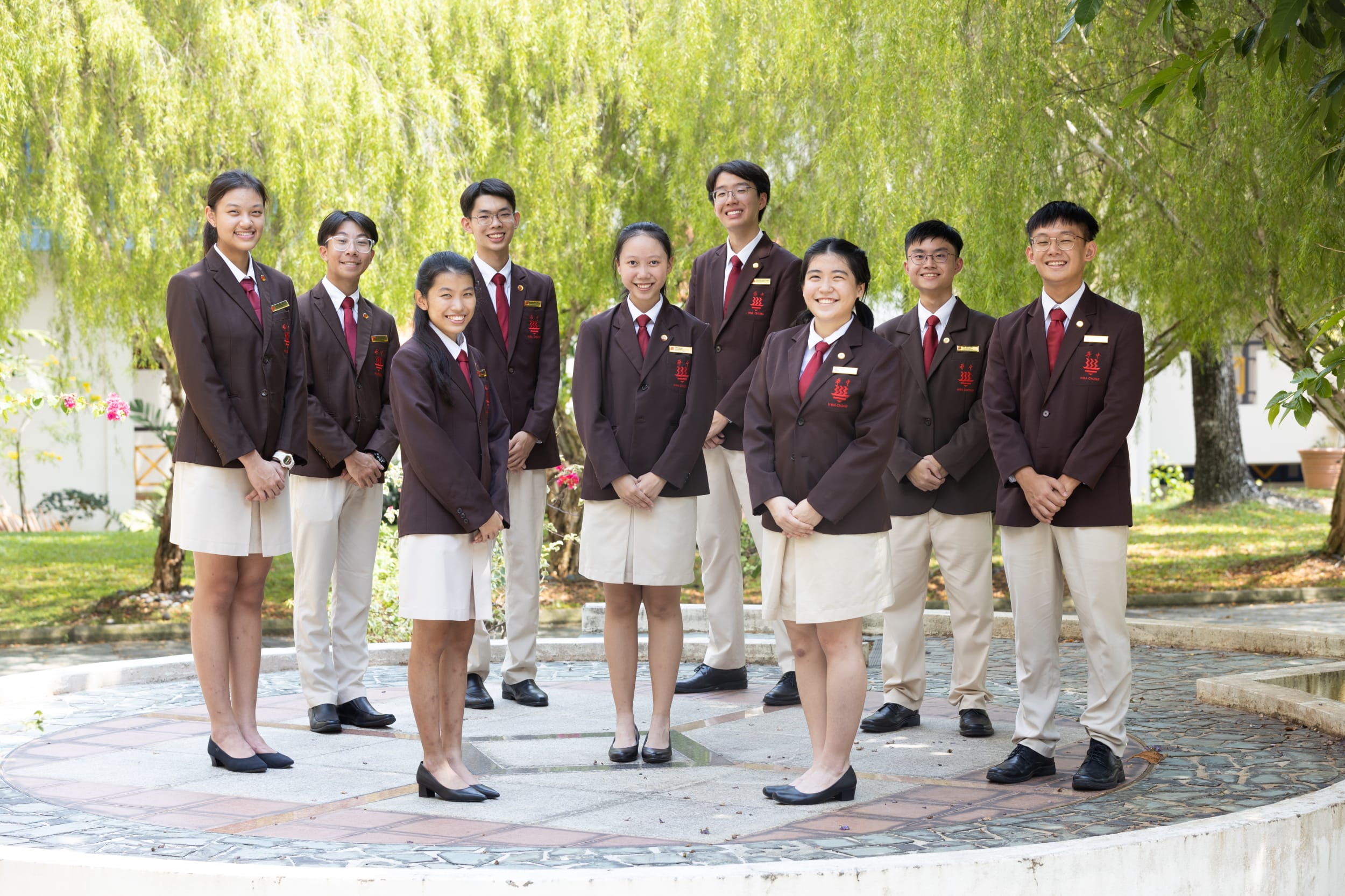 Nine smiling students in matching dark red blazers and red ties pose outdoors on a stone platform near green trees.