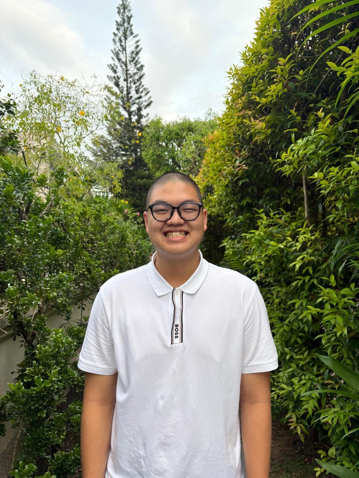 Young man with shaved head and black glasses smiling, wearing a white BOSS polo shirt, standing outdoors among green trees.