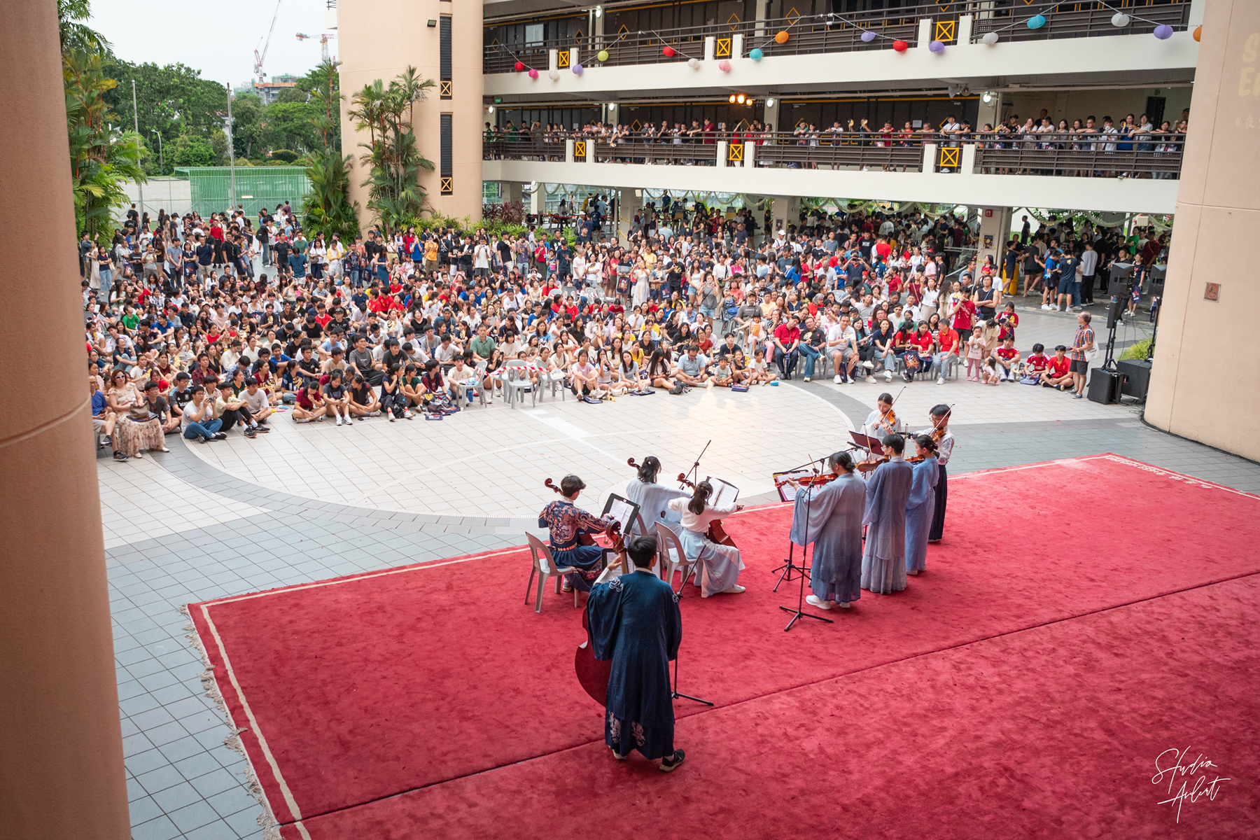 Musicians in flowing costumes perform on a red carpeted stage before a large crowd in a courtyard.