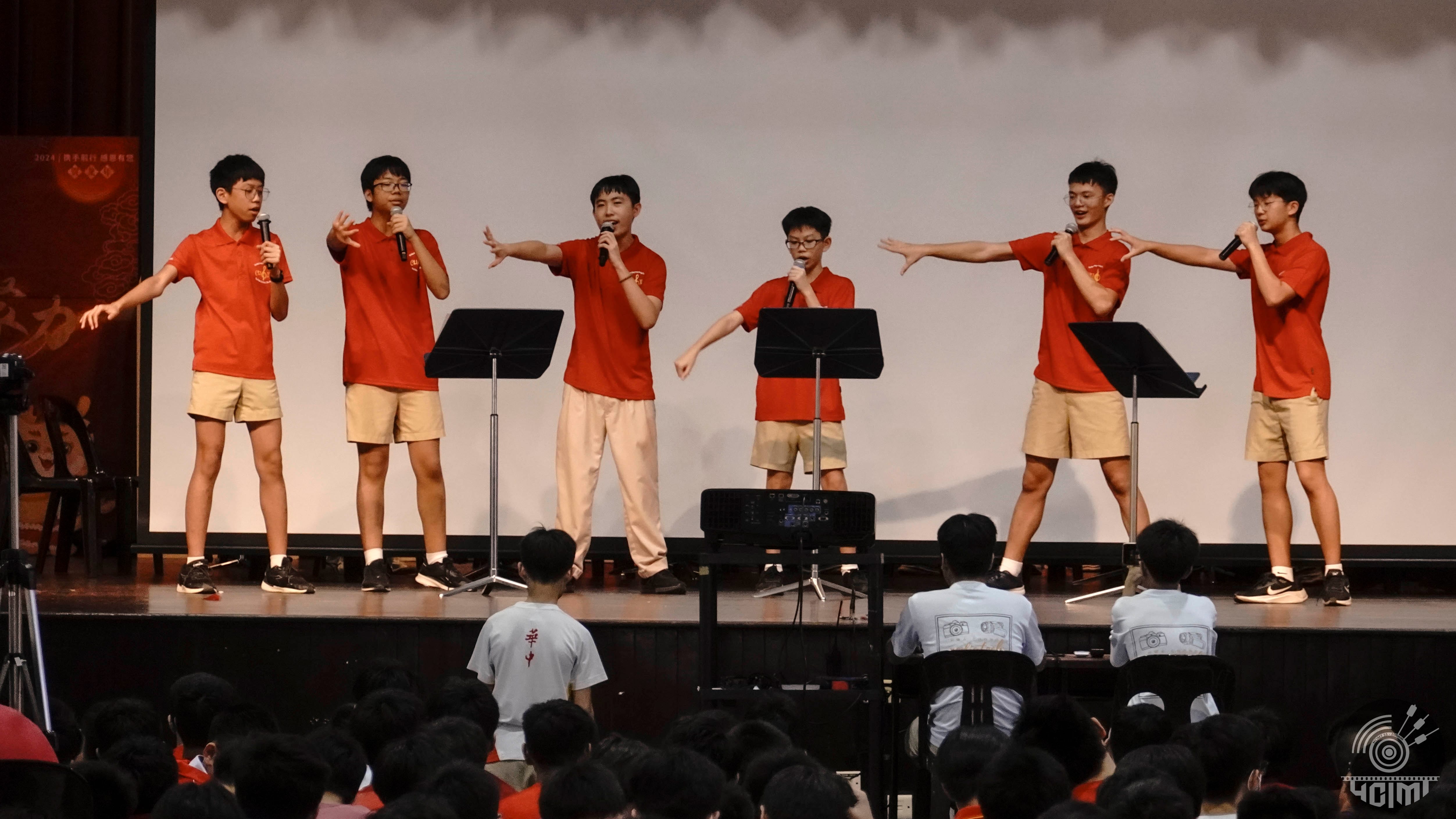 Six boys in red shirts perform on stage with microphones before a seated audience.