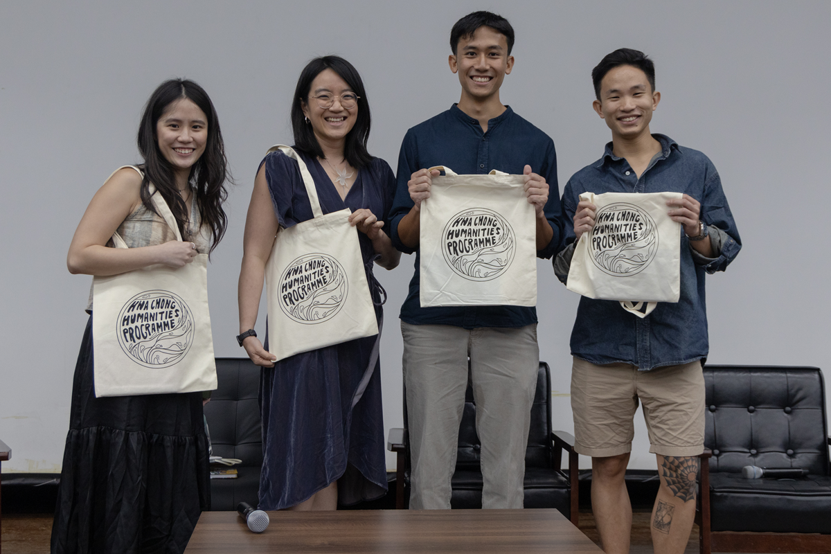 Four smiling people stand together holding cream tote bags printed with Hwa Chong Humanities Programme logo.