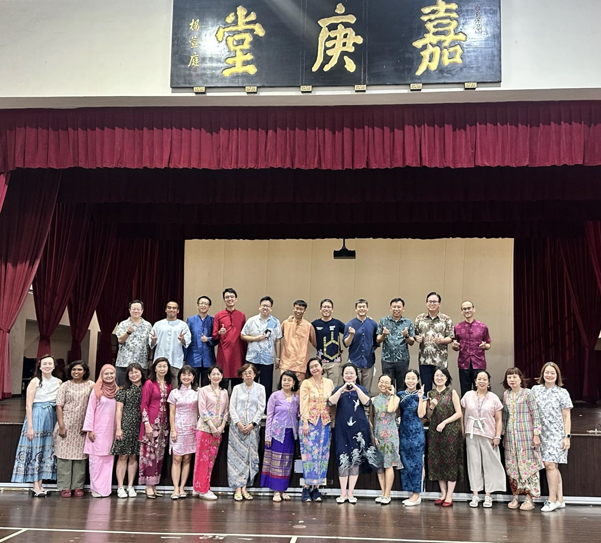A large group photo of school leaders and staff members standing on a stage underneath a traditional signboard at Kah Kee Hall, all dressed in colorful cultural outfits and traditional attire.