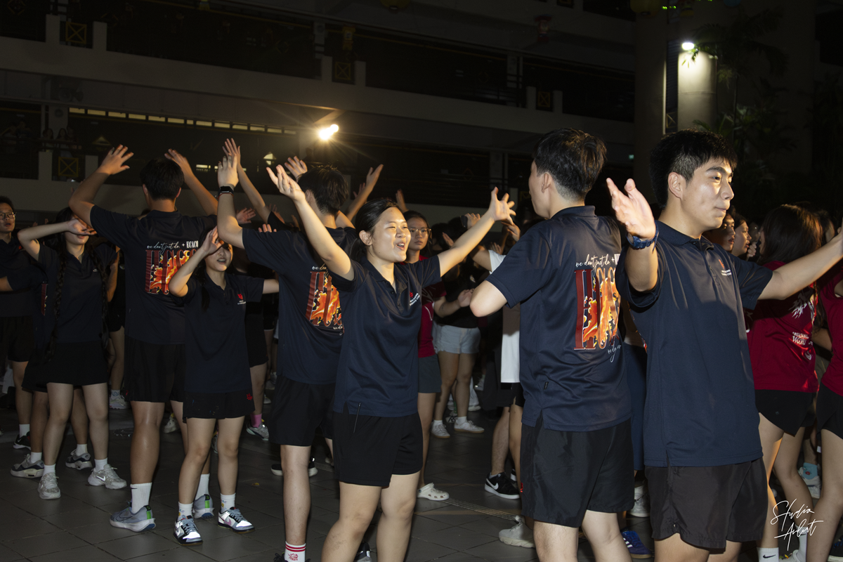 Student councillors wearing dark navy polo shirts dancing with hands raised during an outdoor nighttime campus mass dance session.