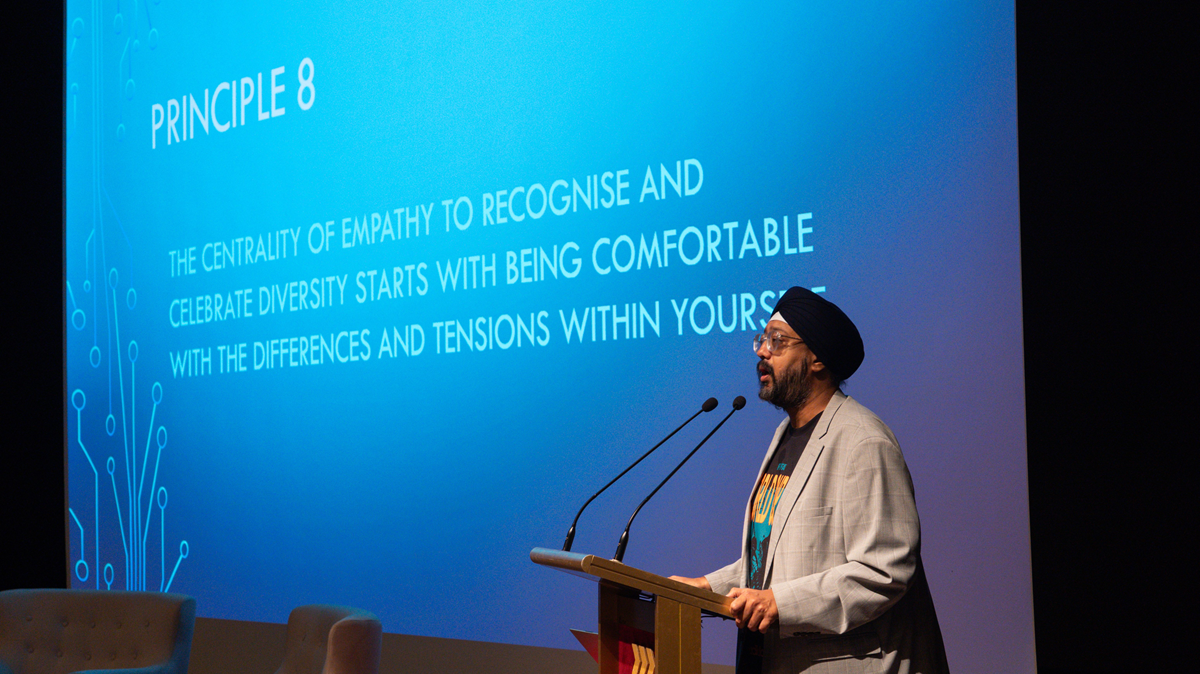 Mr. Malminderjit Singh standing at a podium and delivering a presentation on stage in front of a blue screen that reads "PRINCIPLE 8".