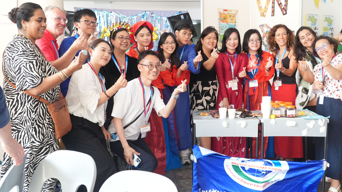 A group of ethnically diverse students and adults smiling, making heart gestures with their hands, and posing next to a cultural display booth.