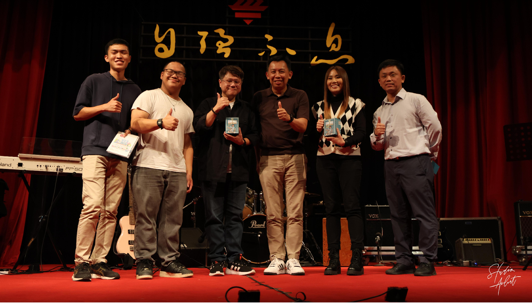 Six people stand on a red stage, smiling and giving thumbs up, with Chinese characters on a dark backdrop.