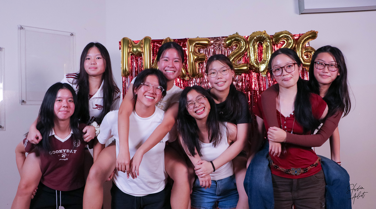 Eight female students posing for a group photo at a photo booth decorated with metallic foil fringe and gold balloon letters spelling MAF 2026.