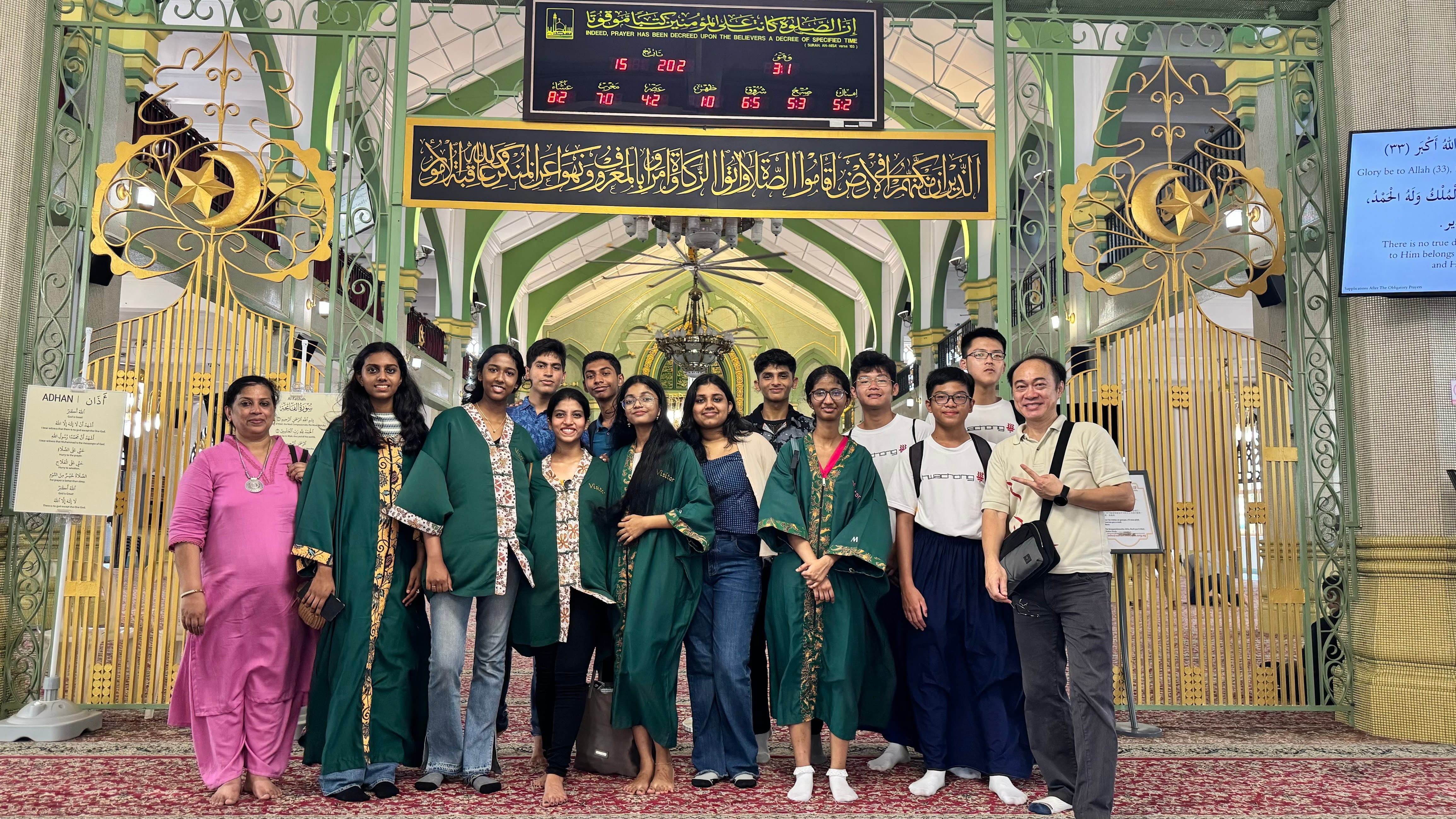 A group of visitors in green robes posing inside a mosque with ornate gold Arabic calligraphy and green archways.