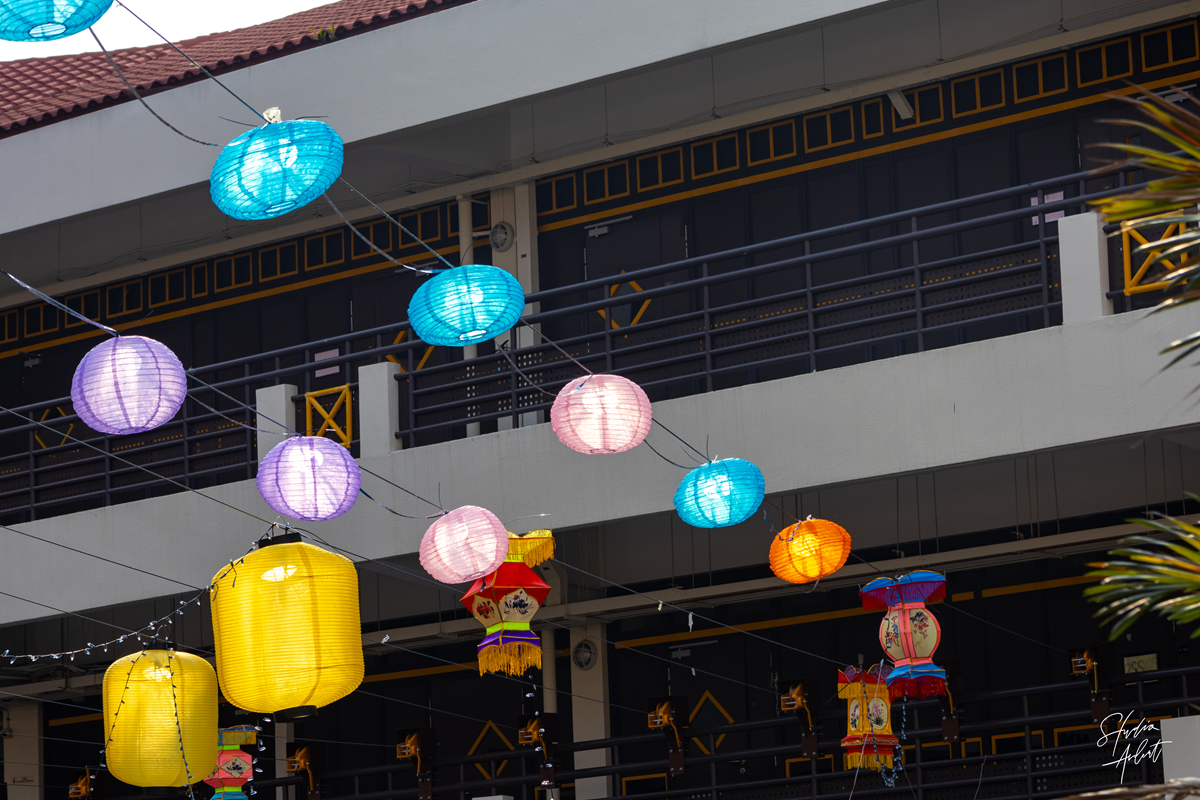 Colorful lanterns in blue, purple, yellow, pink, and orange hanging on strings across a multi-story school campus building for the Mid-Autumn Festival.