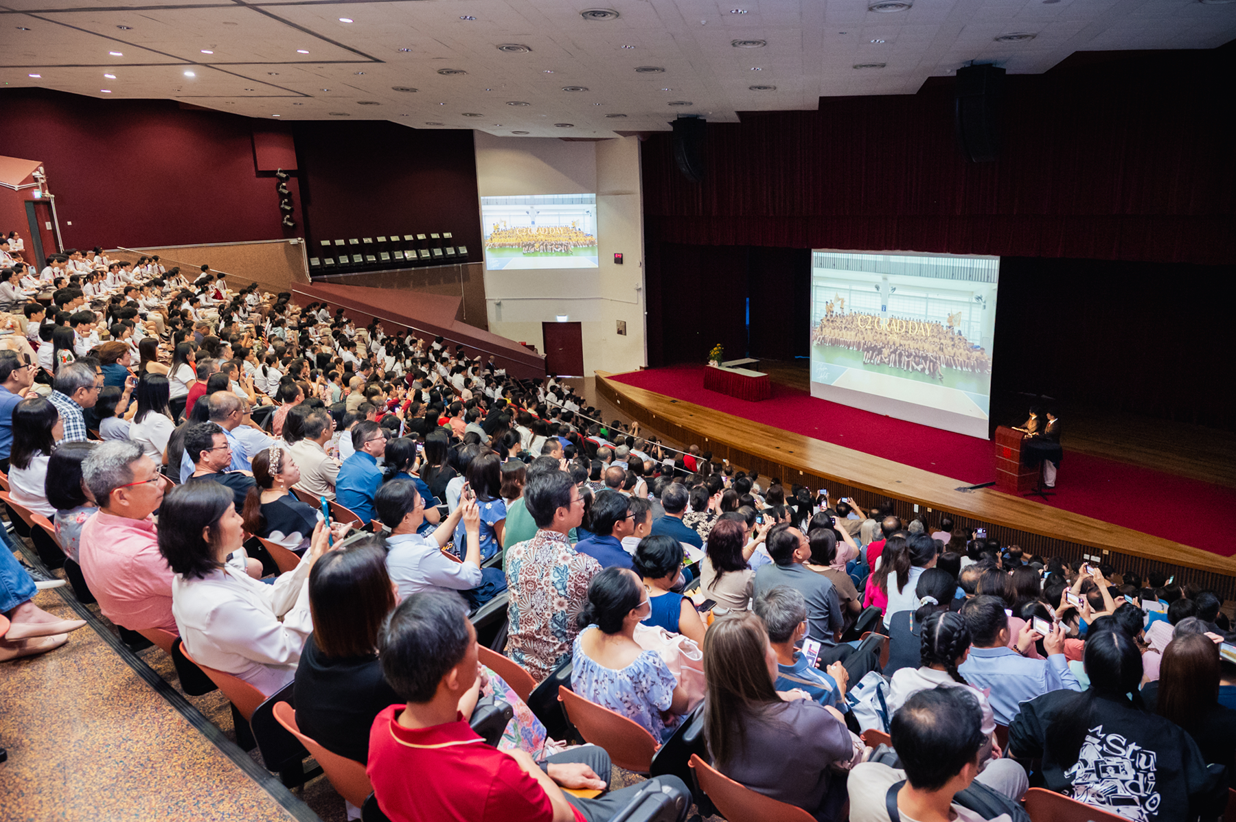 Large auditorium packed with seated audience facing a stage with two big projection screens and a speaker at podium.