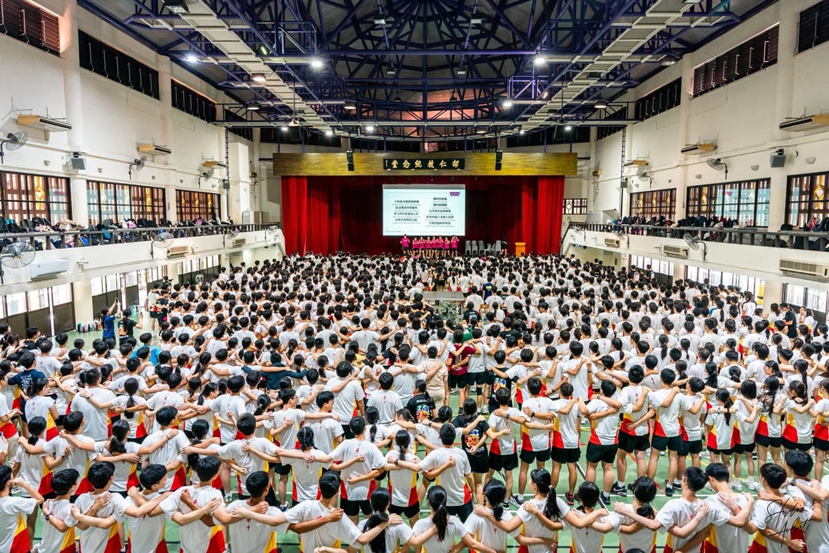 Hundreds of students in white shirts stand linked arm in arm inside a large school hall facing a stage with a screen.