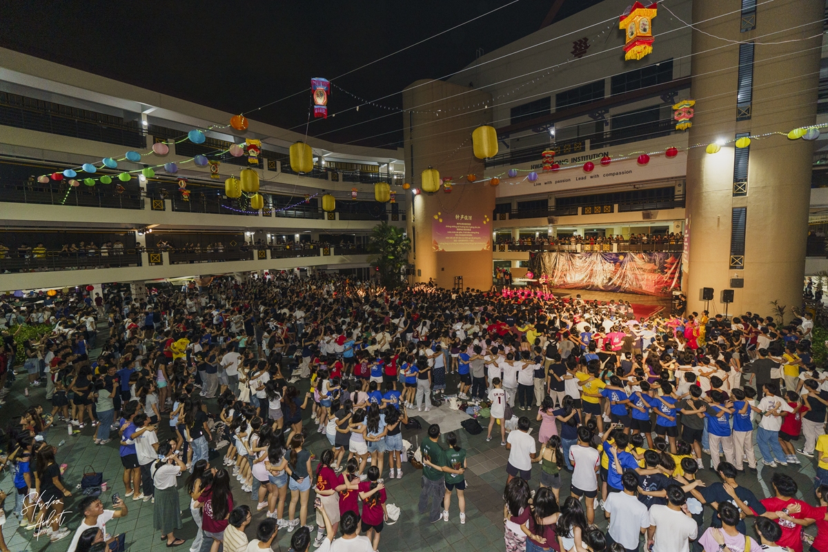 High-angle view of a large crowd of students linking arms and dancing in the central plaza decorated with hanging lanterns during the Mid-Autumn Festival mass SODACHE.