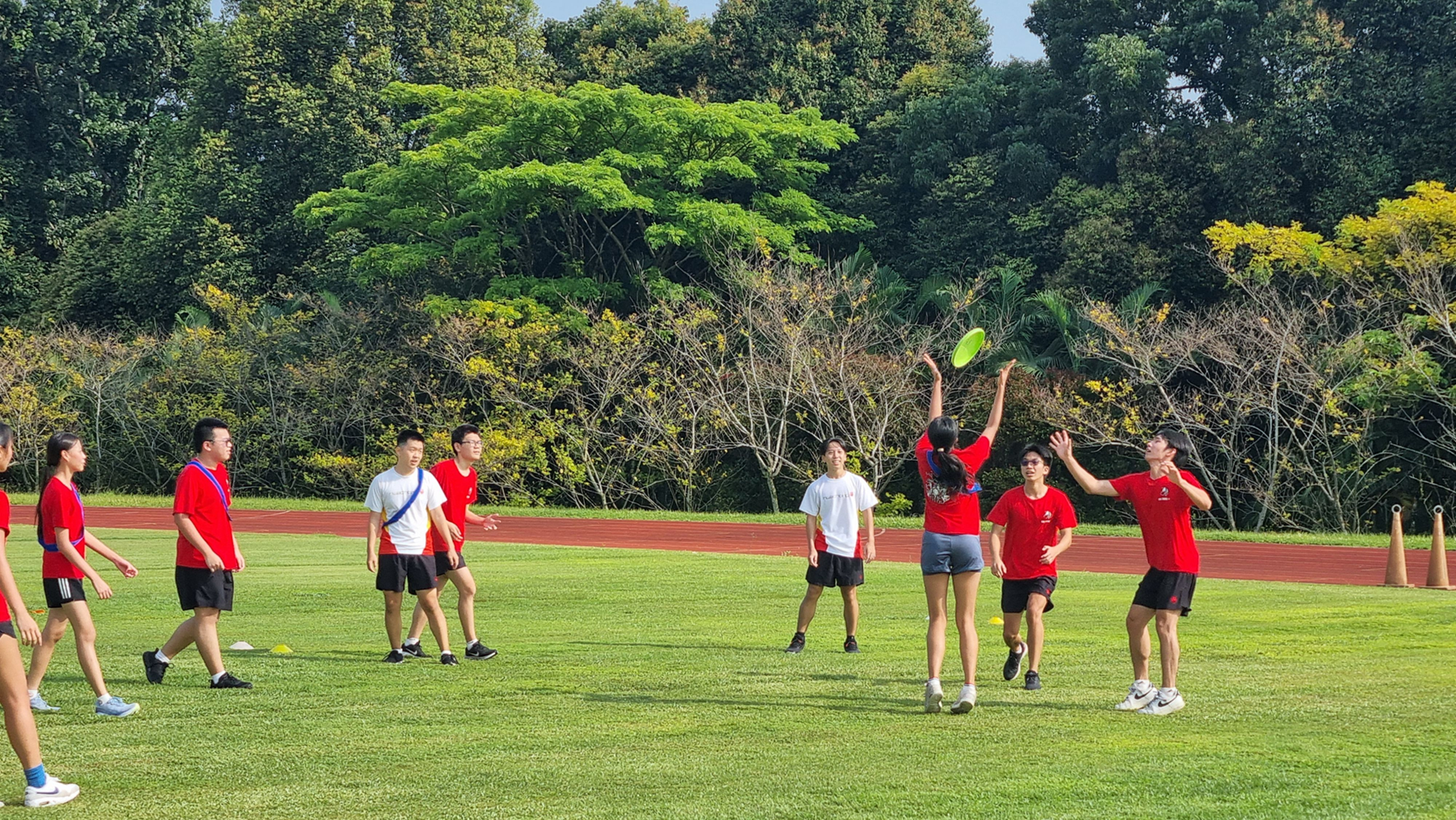 Group in red shirts plays frisbee on a grassy field with a running track and trees behind.