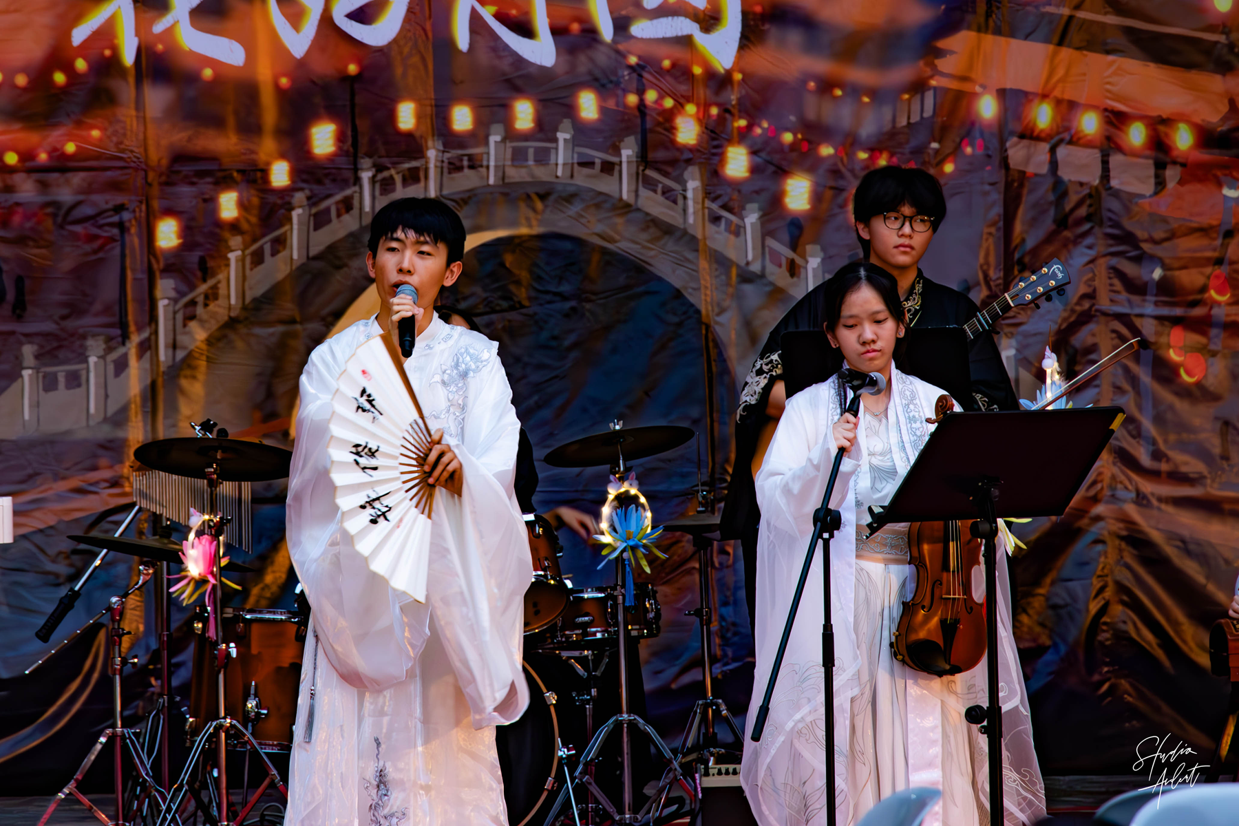 Three young performers on stage in white and black hanfu, one singing with a fan, one holding a violin.
