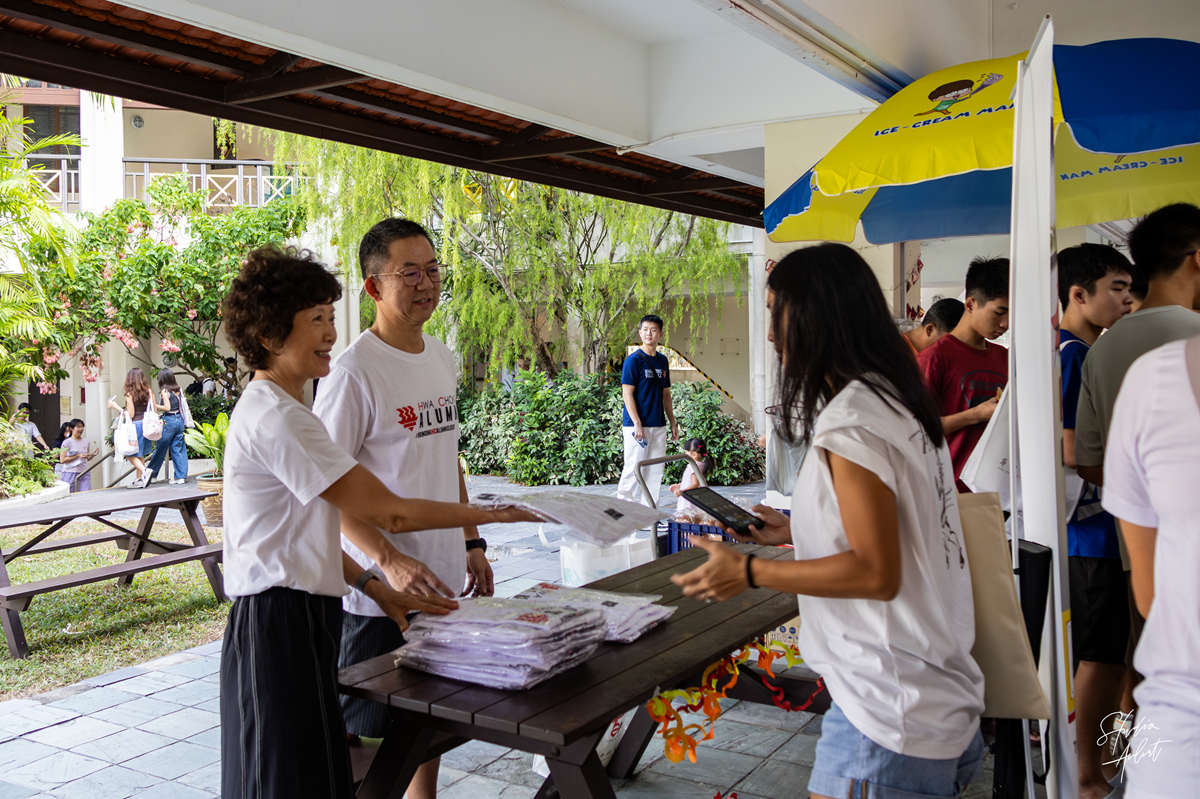 Alumni volunteers (HCAA) in white shirts handing out commemorative shirts to a visitor at an outdoor collection table.
