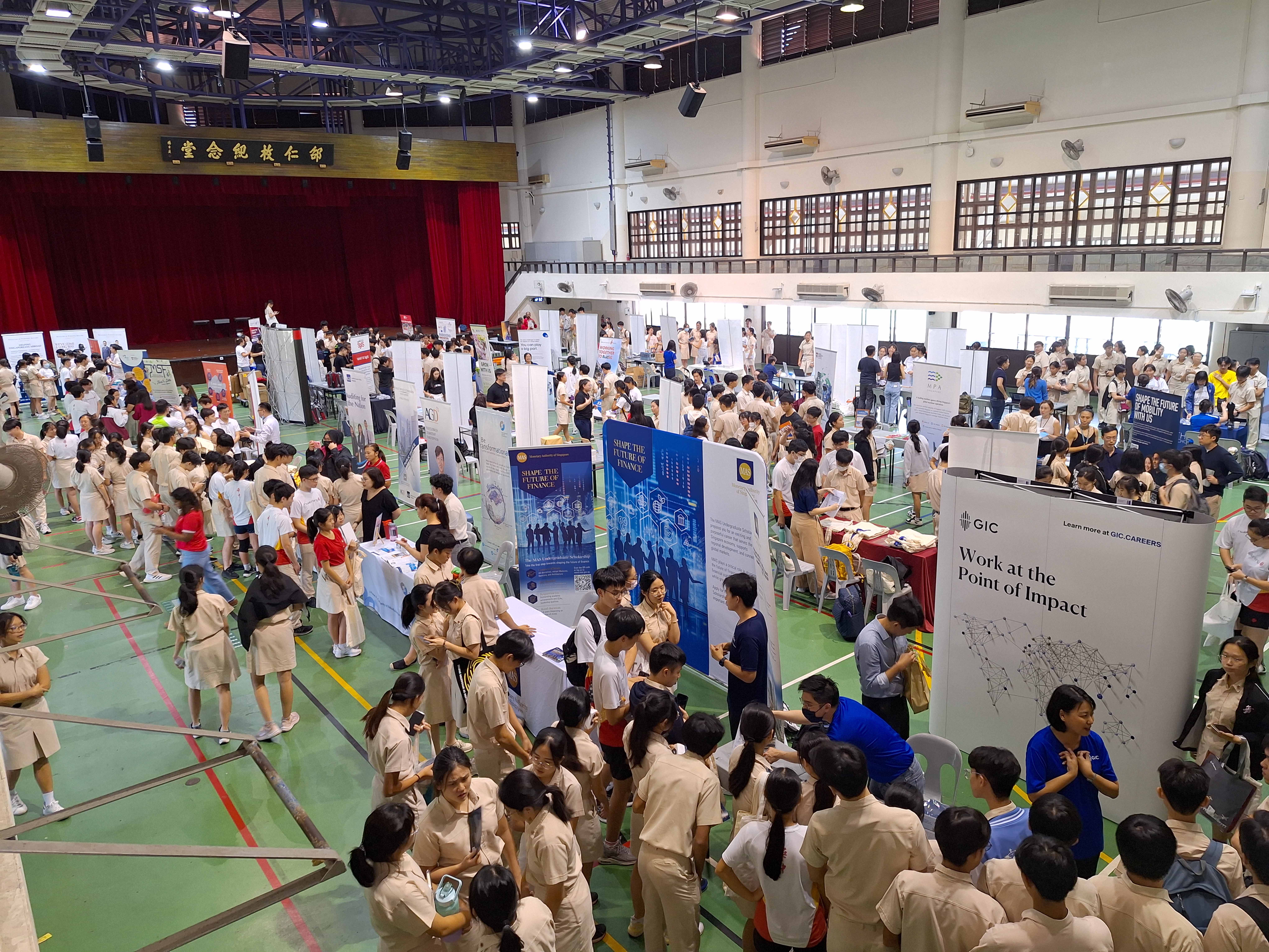Crowded indoor school fair with students in uniforms around booth displays, near a stage with red curtains.