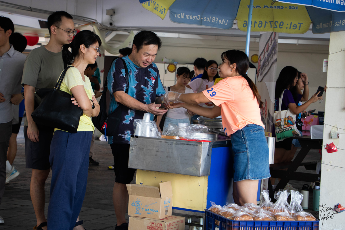 A stall vendor in a peach shirt handing a treat from an ice cream cart from HCAA to visitors at an outdoor festival booth.