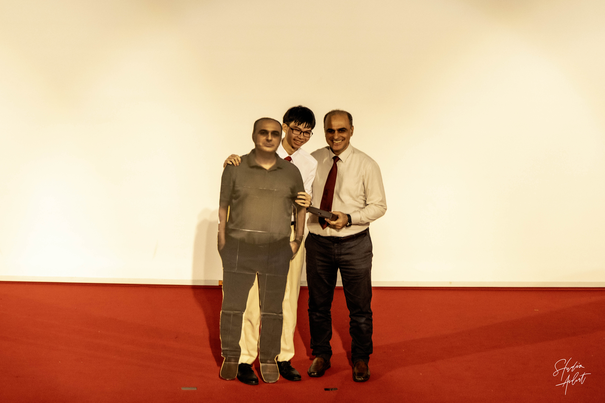 Two men and a life-size cardboard cutout of a person standing together on a red floor against a cream wall.