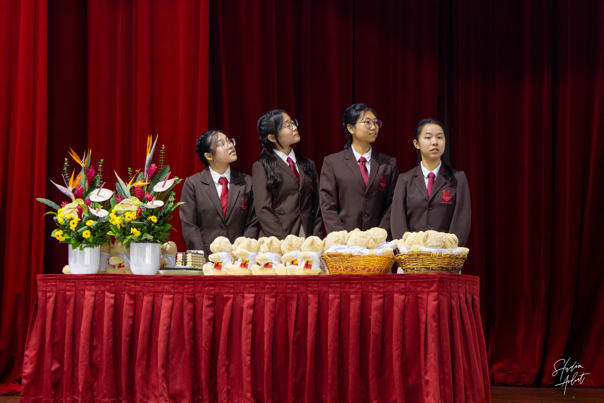 Four students in brown blazers and red ties stand behind a table of bread, baskets, and flowers on red stage curtains.