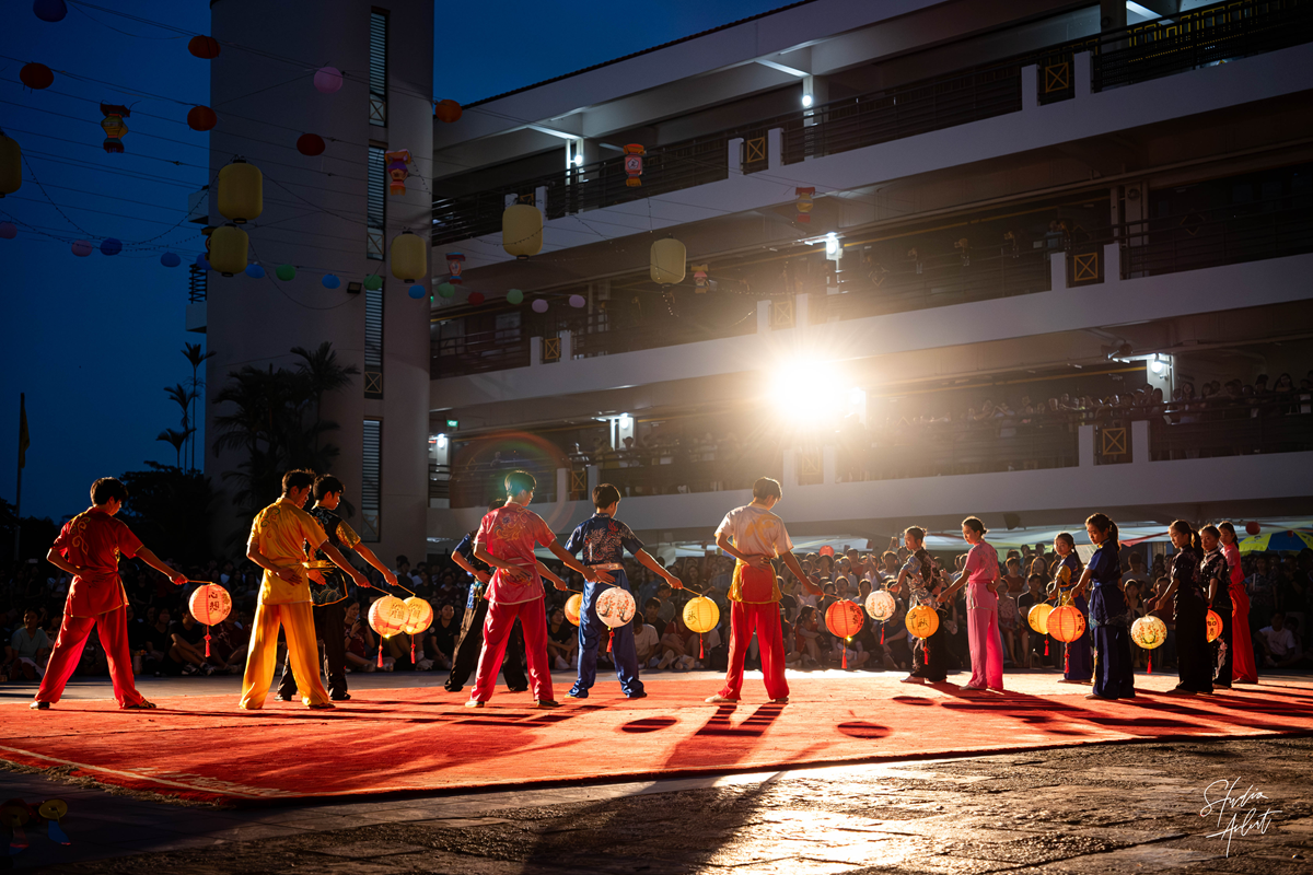 Student wushu performers in colorful traditional costumes holding glowing paper lanterns during a nighttime outdoor stage showcase.