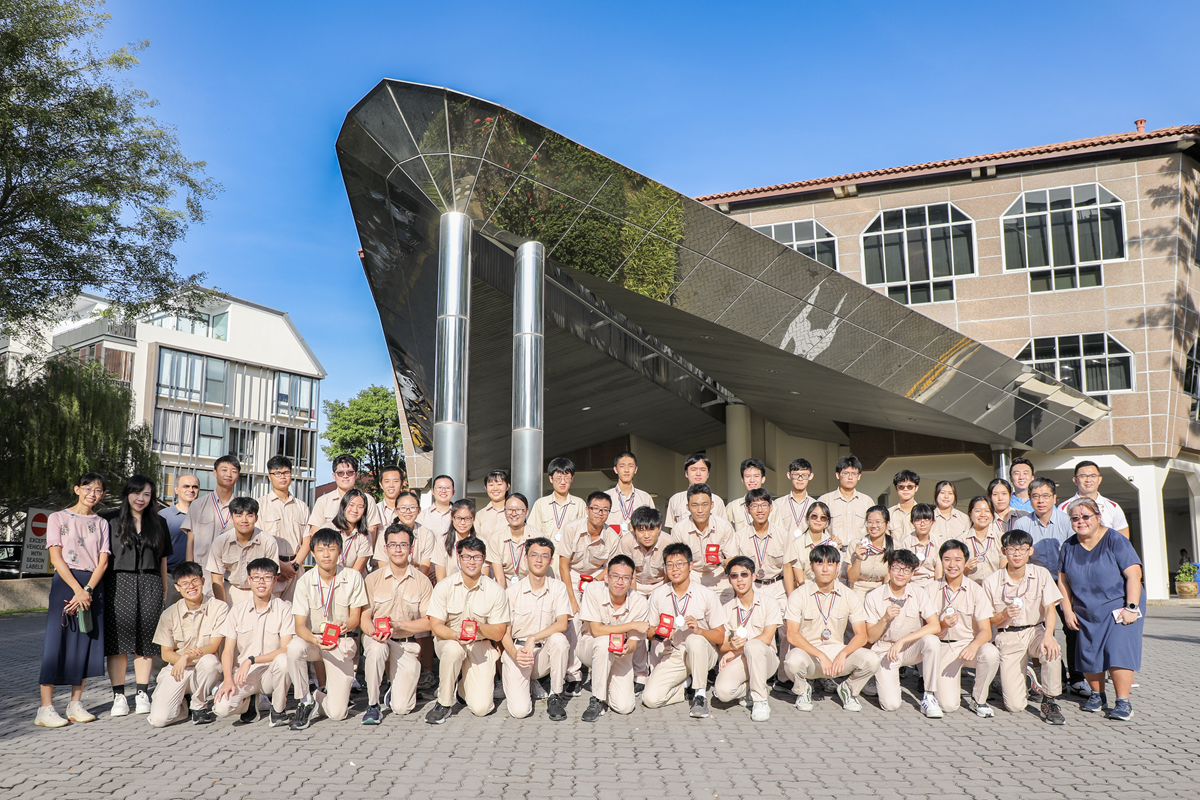Large group of students in beige uniforms posing with medals in front of a modern reflective building outdoors.
