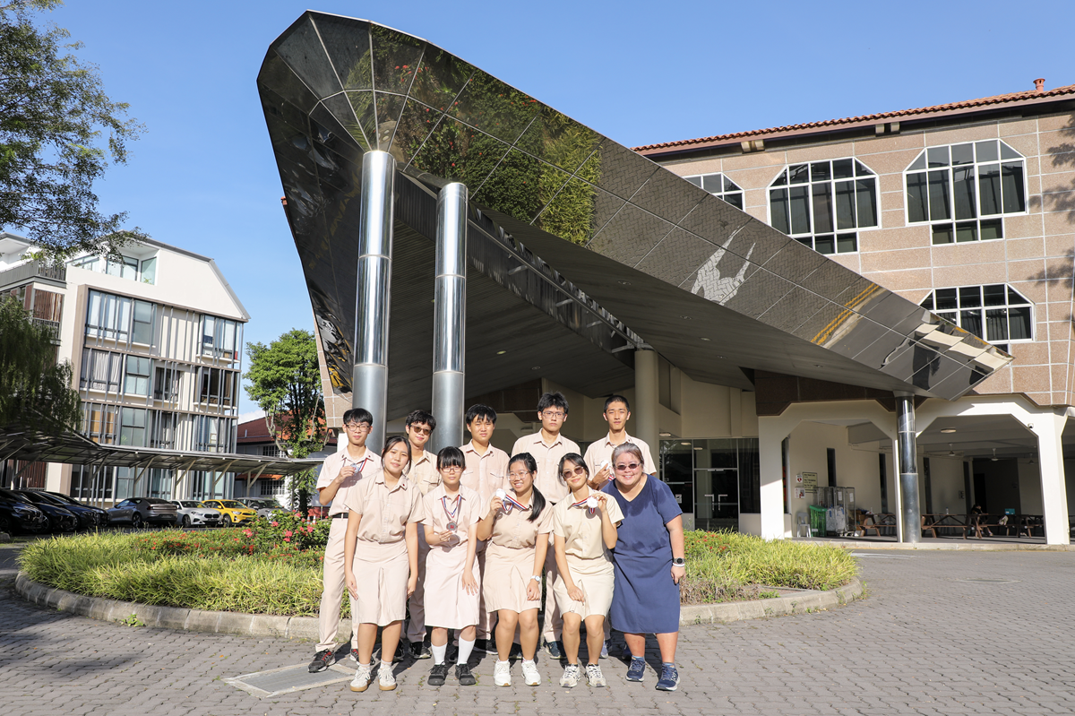 A group of students in beige uniforms and an adult in blue pose outside a school building with a modern canopy.