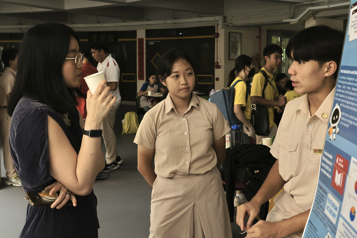 Three young people talk near a blue presentation board in a busy school corridor, with students in the background.