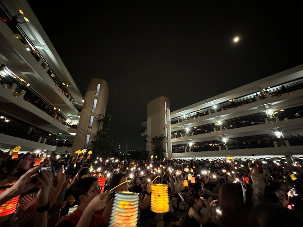 Large crowd holding colorful lanterns and phone lights at night between multi-story buildings under a moonlit sky.