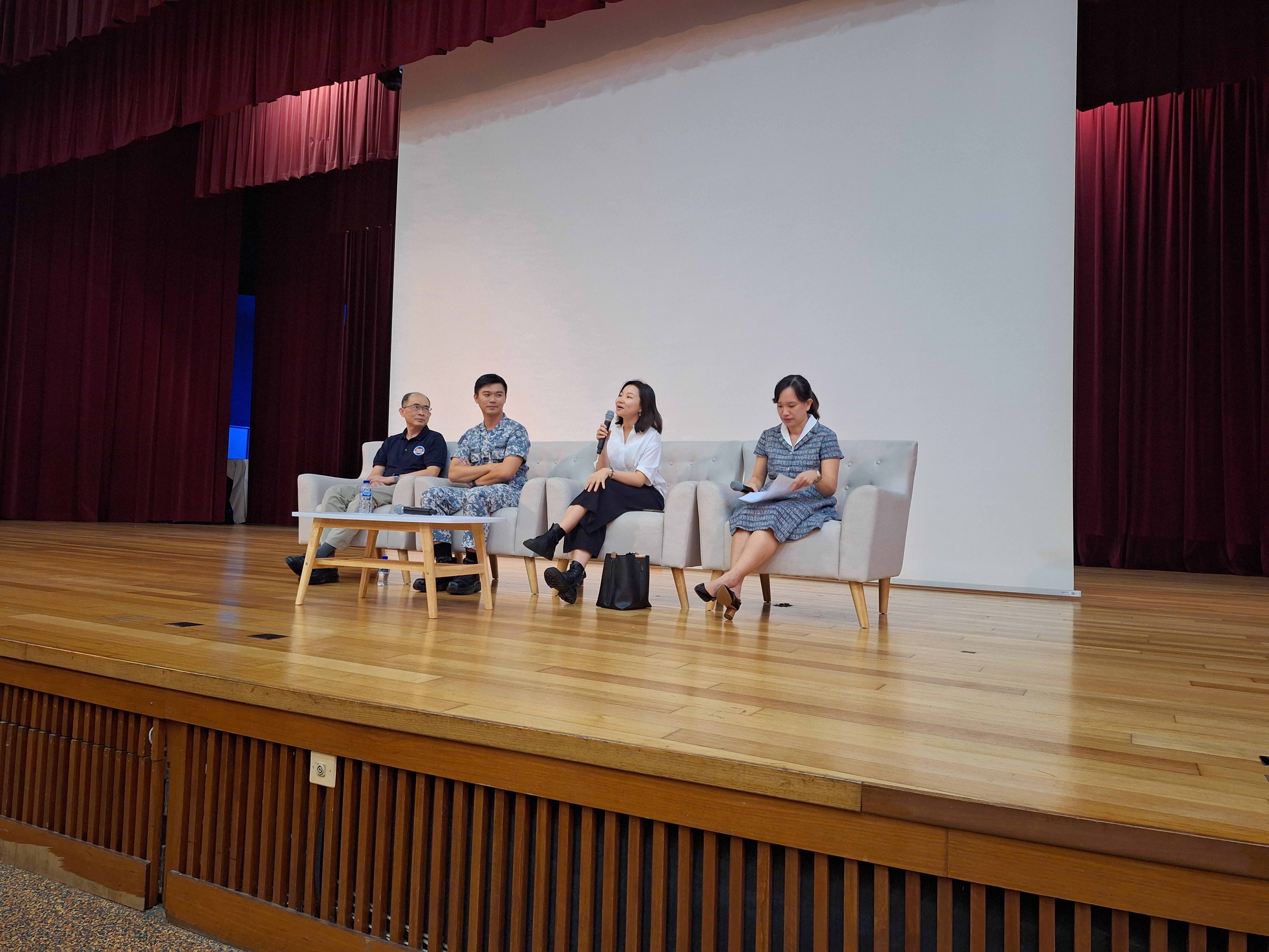 Four panelists on sofas on a stage; one woman speaks into a microphone, another reads notes.