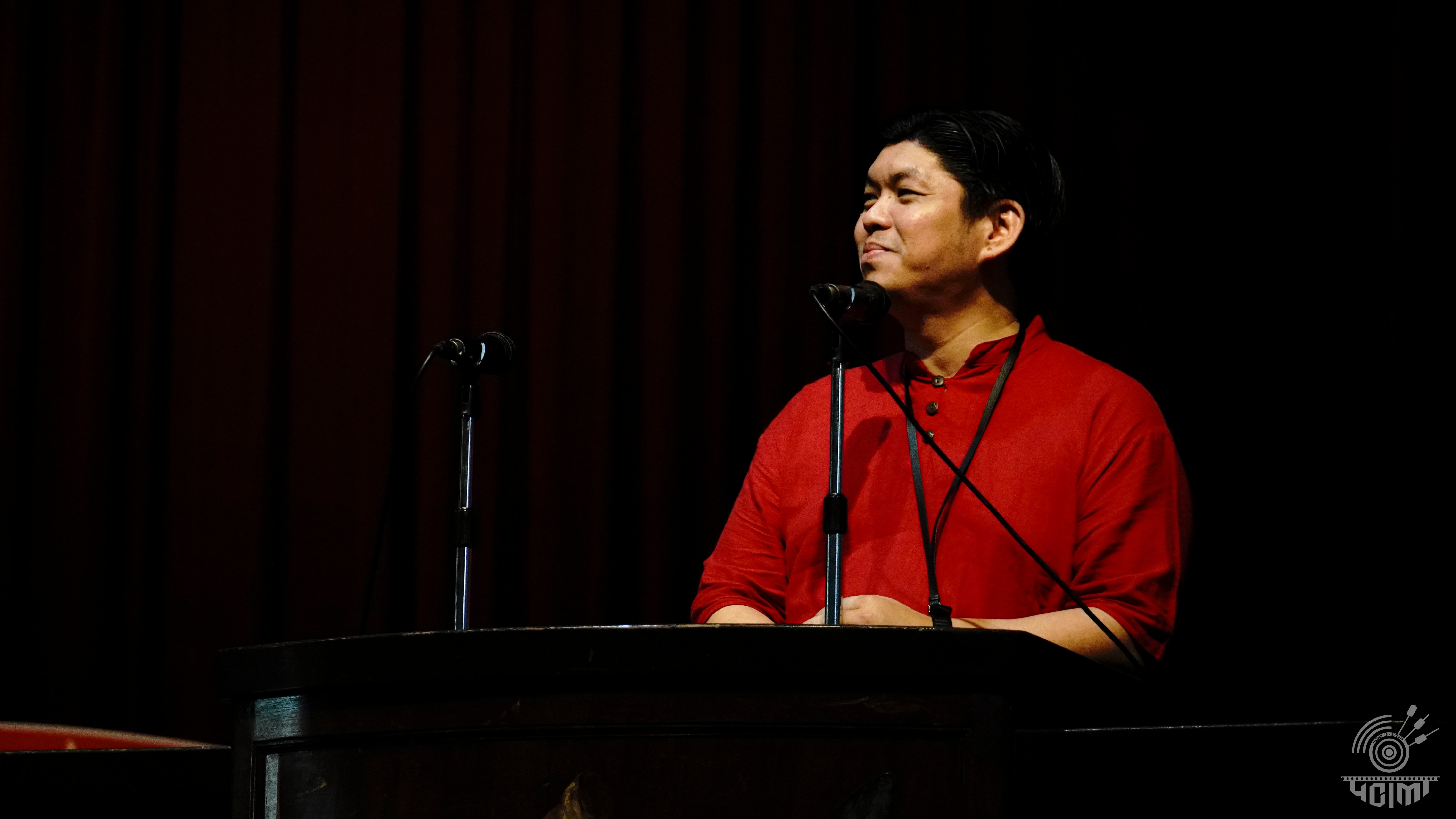 Man in a red shirt with a lanyard stands at a dark podium with two microphones, looking to the side.