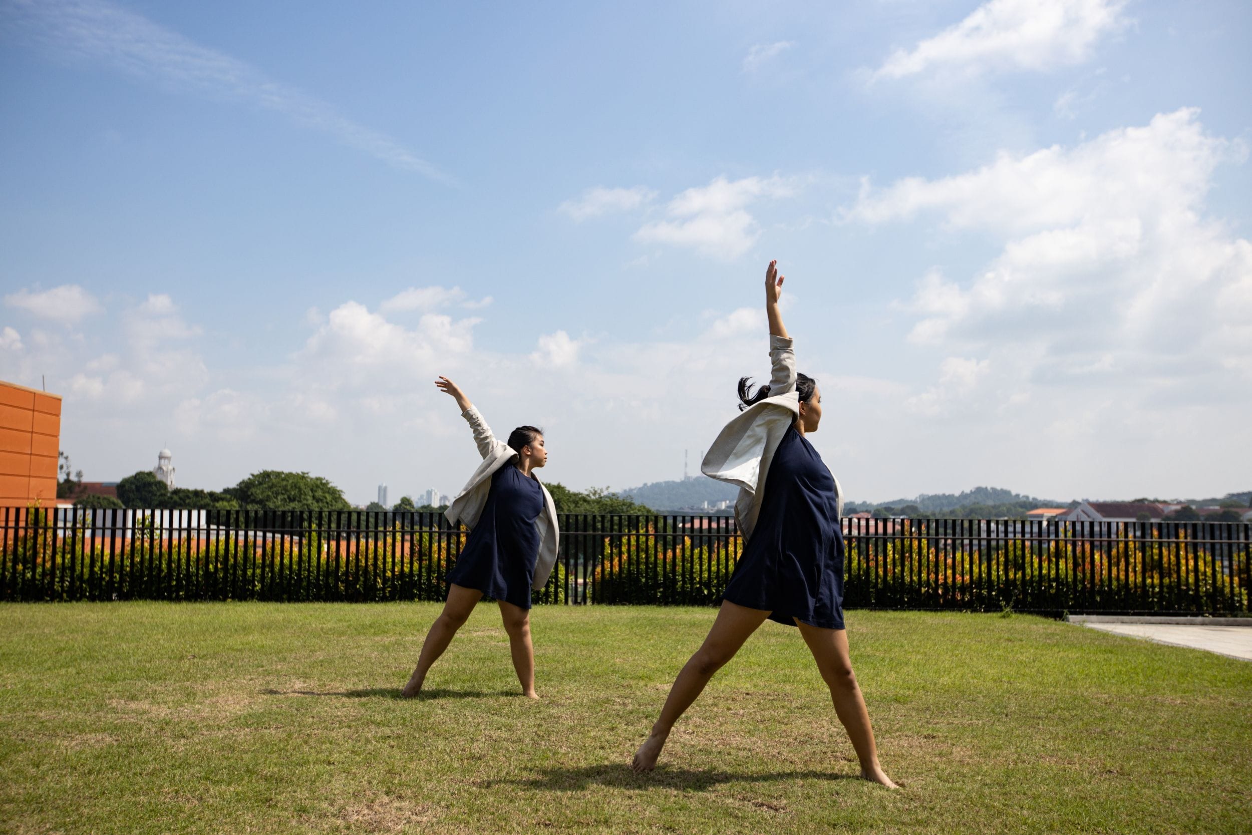 Two barefoot dancers in navy outfits stretch arms upward on a grassy rooftop under a partly cloudy sky.