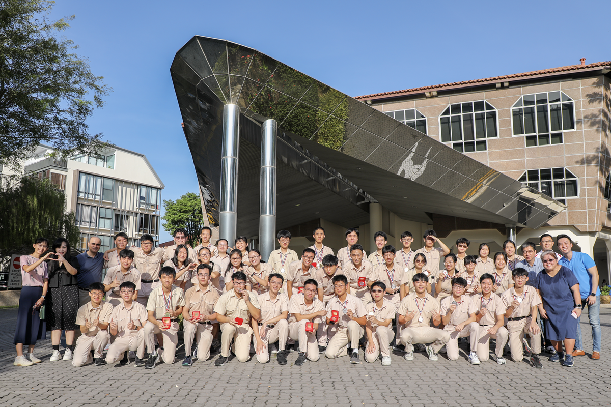 A large group of students in tan uniforms holding medals pose outdoors in front of a modern angular building.
