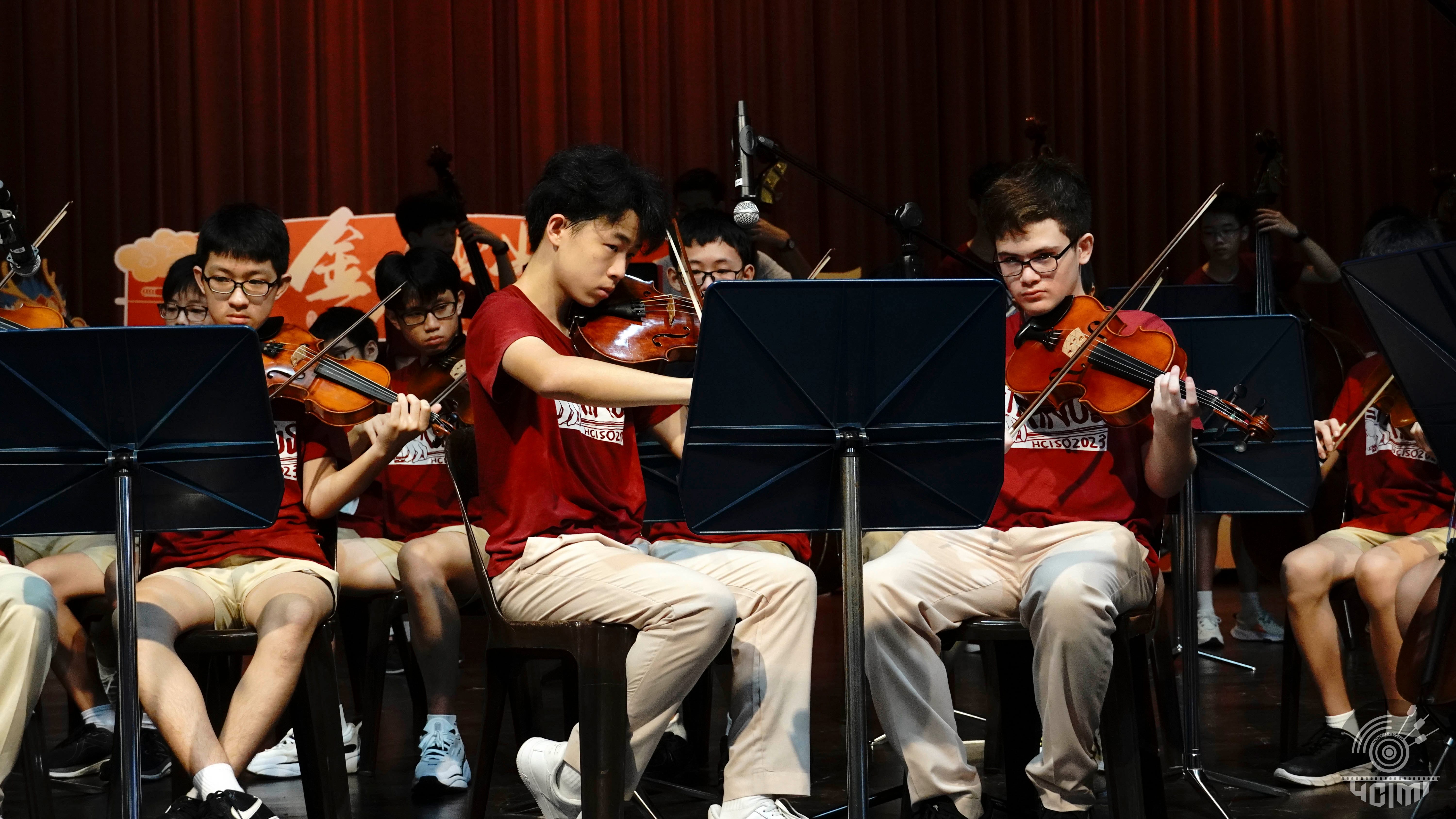 Young students in red shirts play violins on stage, seated before music stands with a red curtain behind them.
