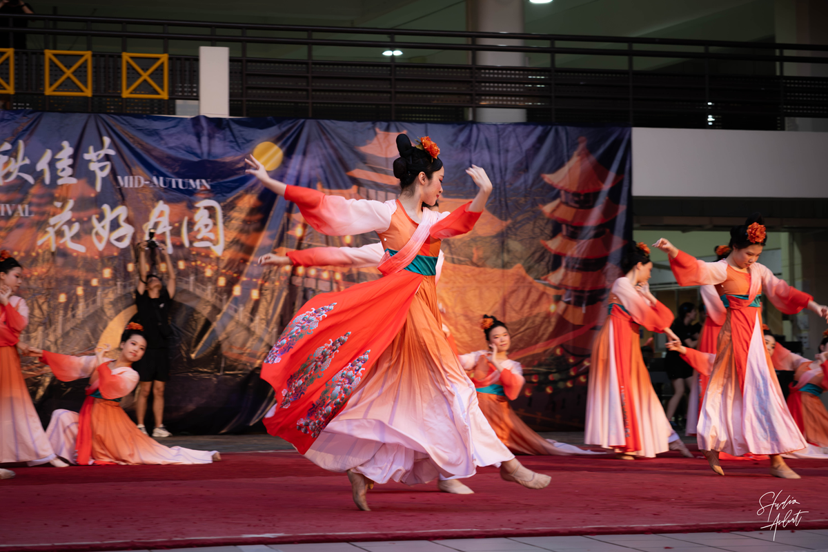 Student dancers performing a Chinese dance in flowing red and orange traditional costumes on stage during the Mid-Autumn Festival.
