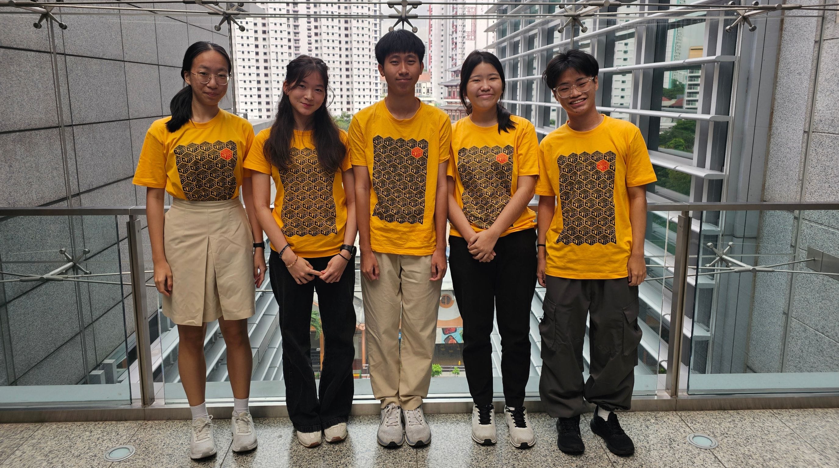 Five young people wearing matching yellow patterned t-shirts stand together on an indoor balcony with city buildings behind them.
