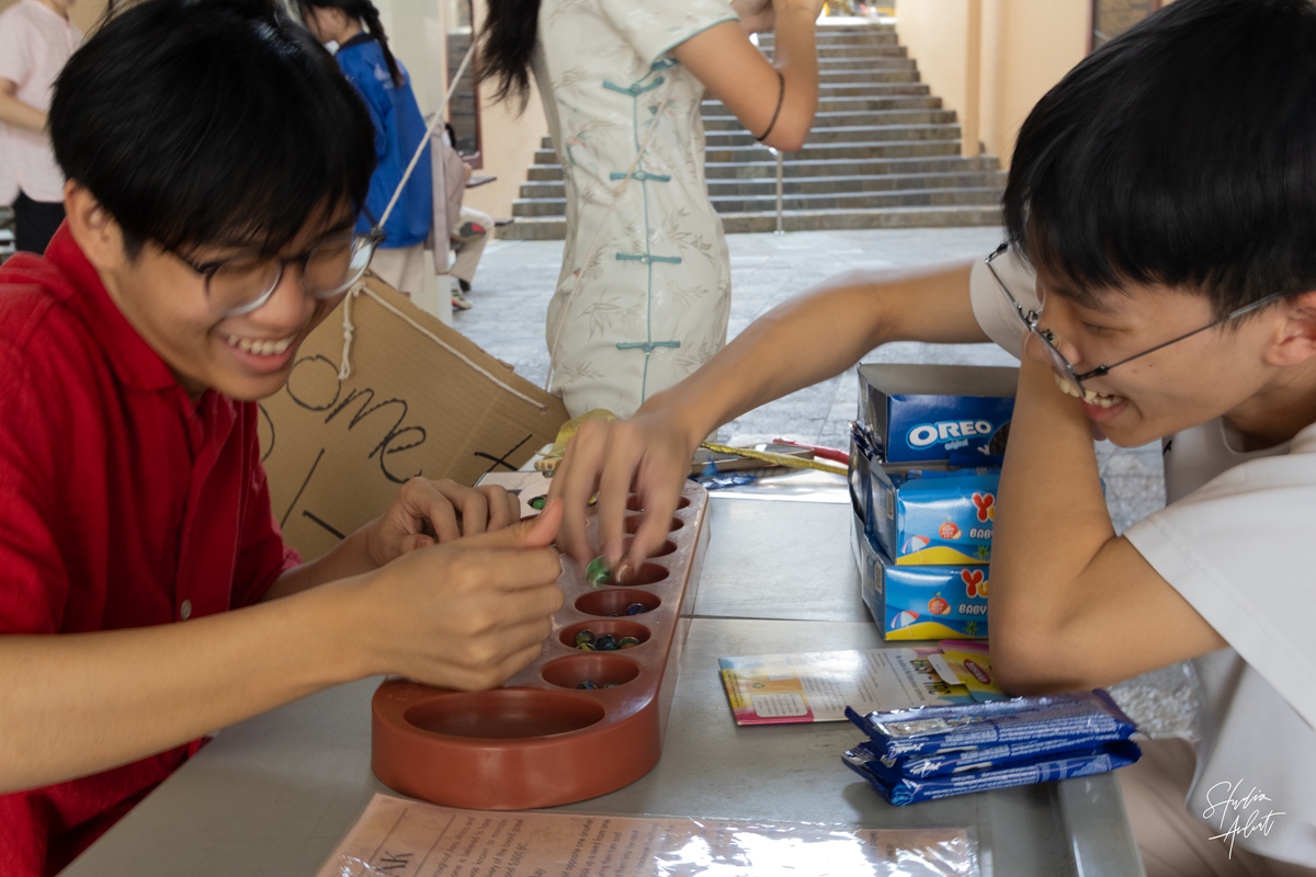 Two male students smiling and laughing as they play the traditional game of Congkak at a booth setup during the school event.