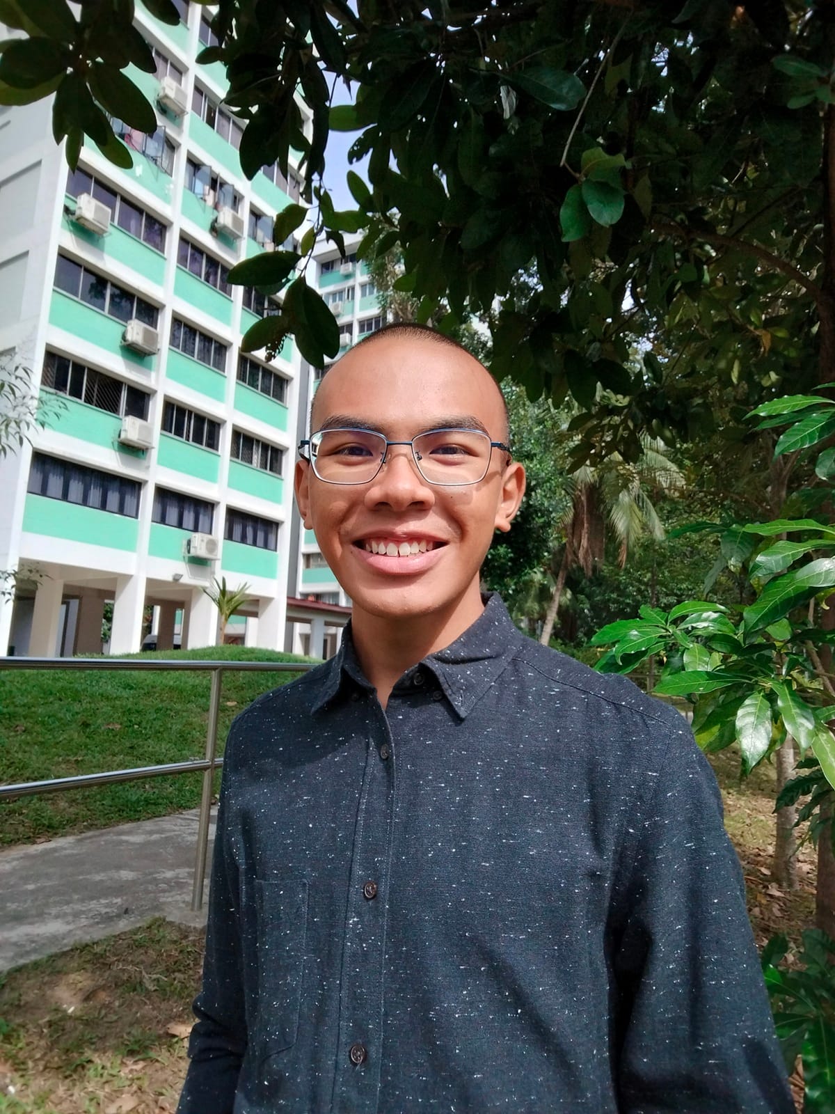 Young man with shaved head and glasses smiling outdoors, wearing a dark shirt, with a green building and trees behind him.