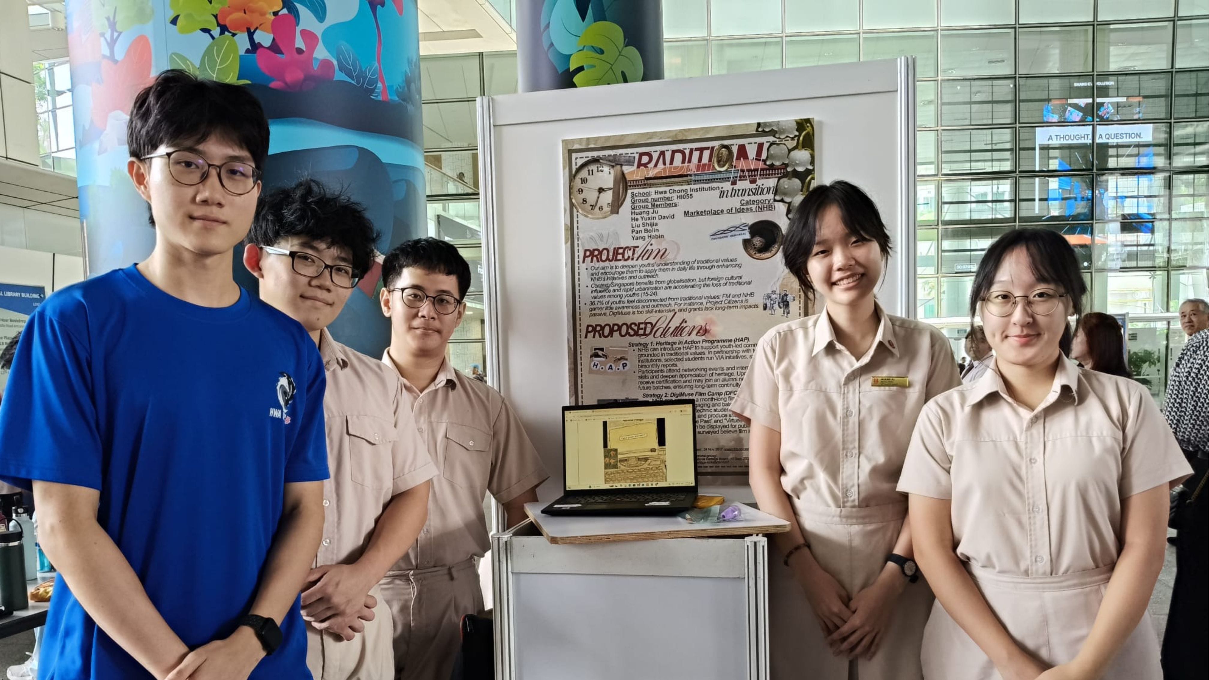 Five students in school uniforms stand beside a project display board and laptop at an indoor exhibition.