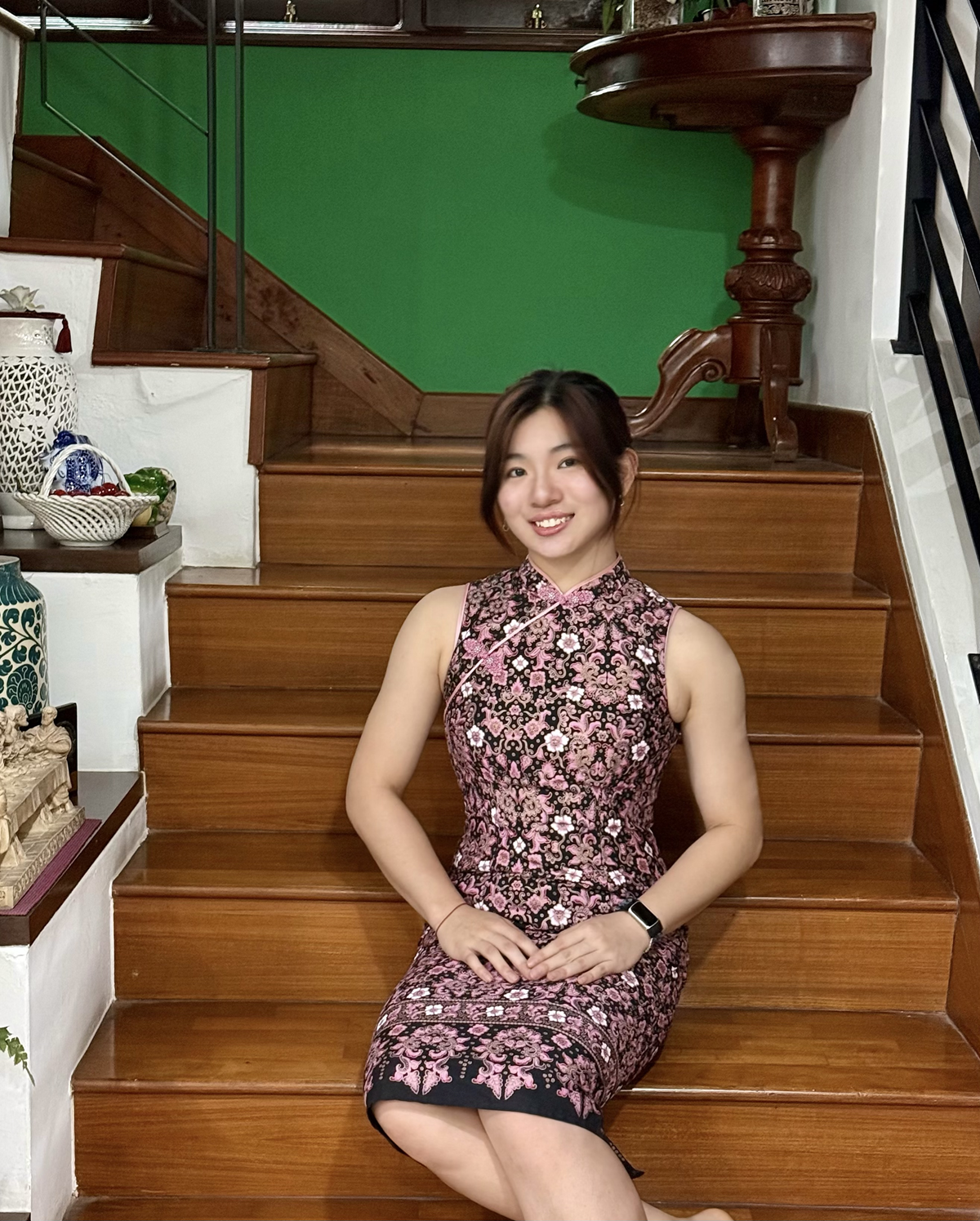 Young woman in a black and pink floral cheongsam dress sits smiling on wooden indoor stairs.
