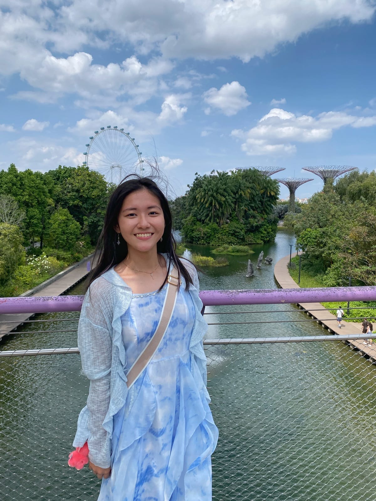 Young woman in a blue dress smiling on a bridge at Gardens by the Bay, Singapore, with Supertrees and a Ferris wheel behind her.