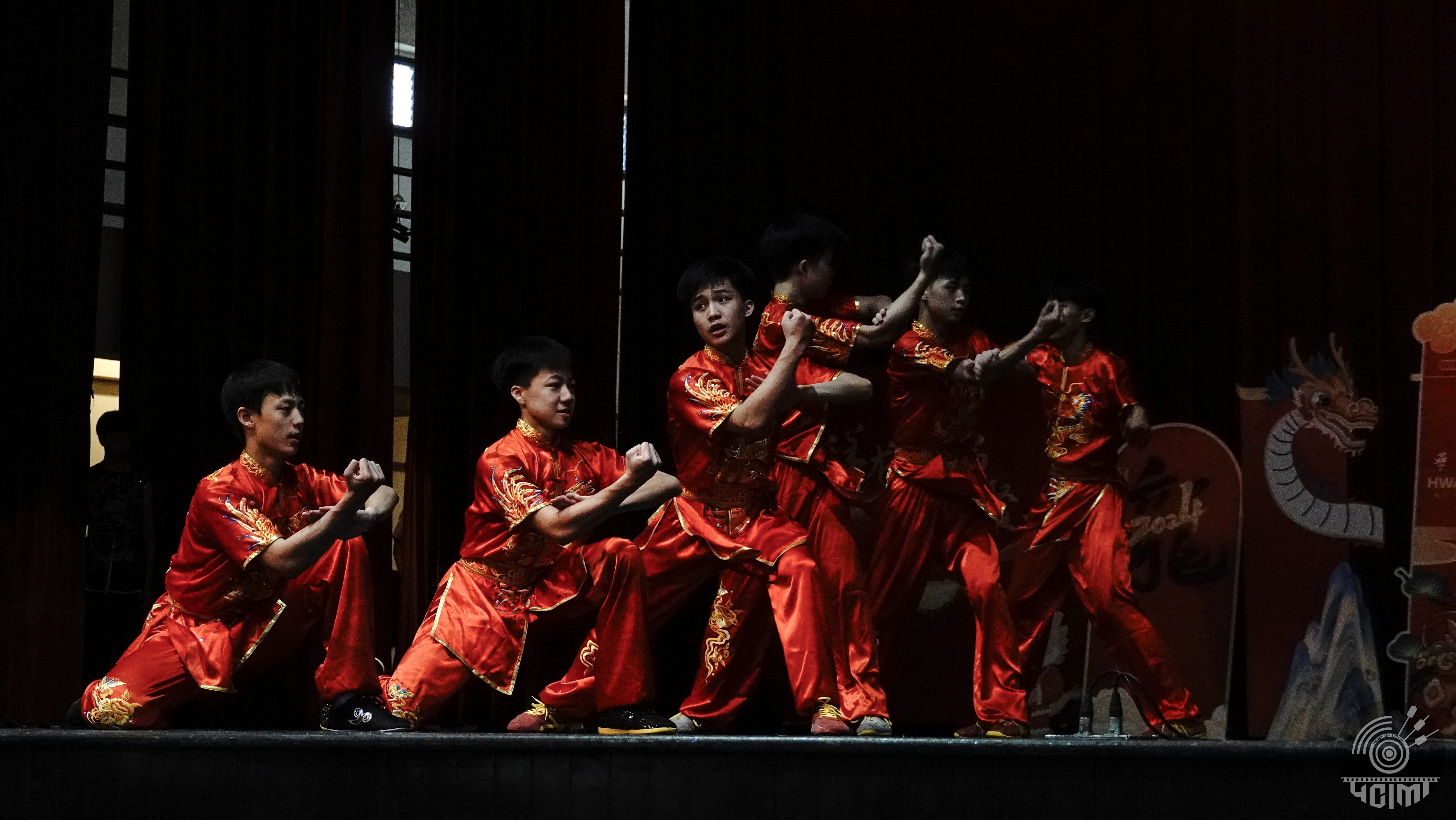 Several young performers in red and gold embroidered wushu uniforms strike martial arts poses on a dark stage.
