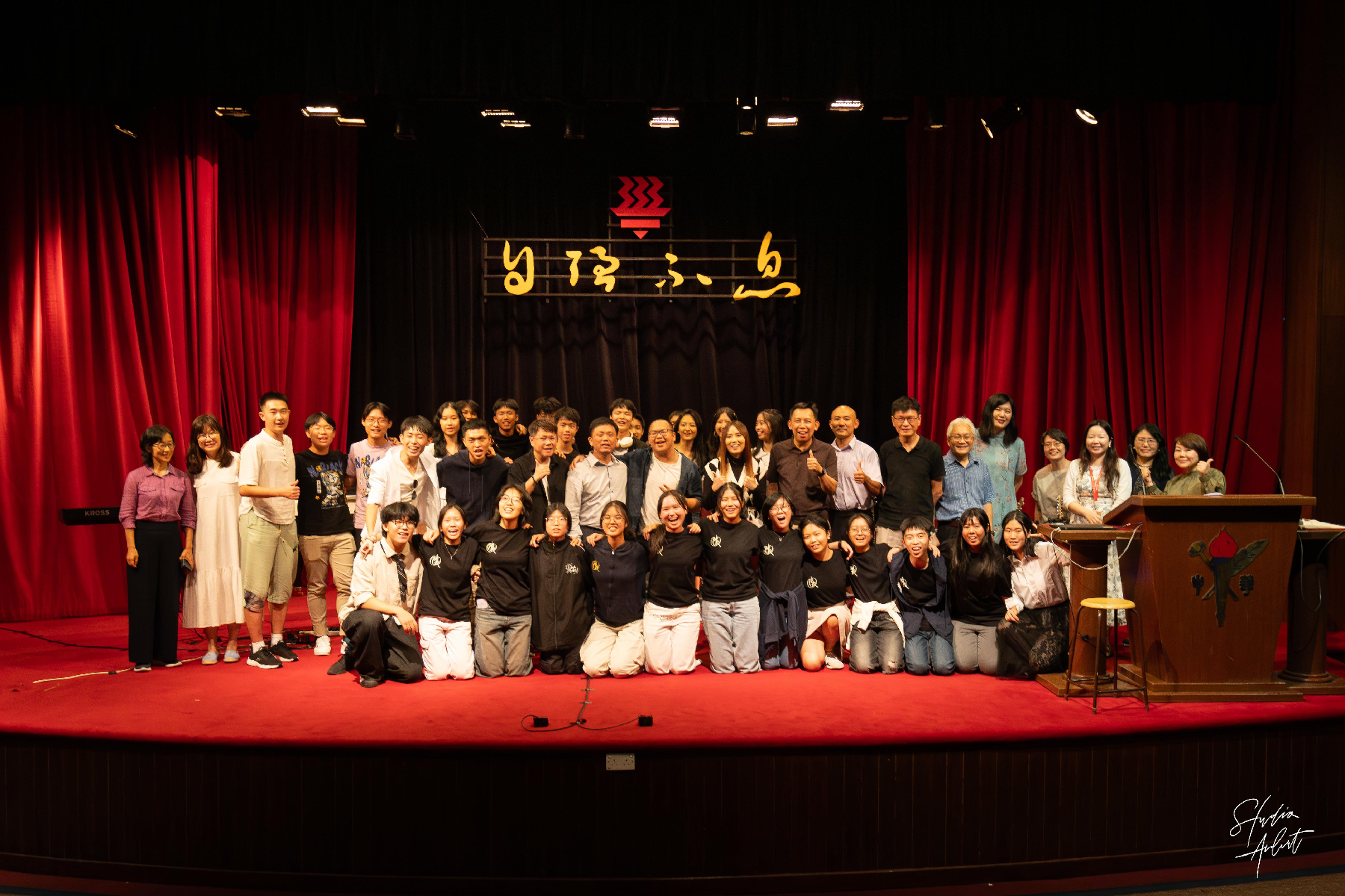 Large group of people posing on a red stage with red curtains and a Chinese character sign behind them.