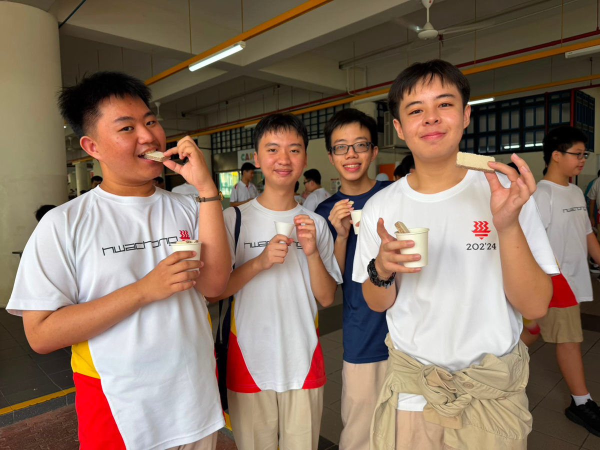 Four male high school students in white school shirts smiling and posing for the camera while holding up cup corn, ice cream, and retro wafer biscuits at the canteen booths.