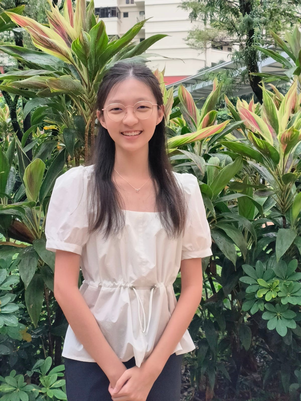 Young woman with long dark hair and glasses, smiling, wearing a white top, standing in front of tropical plants.